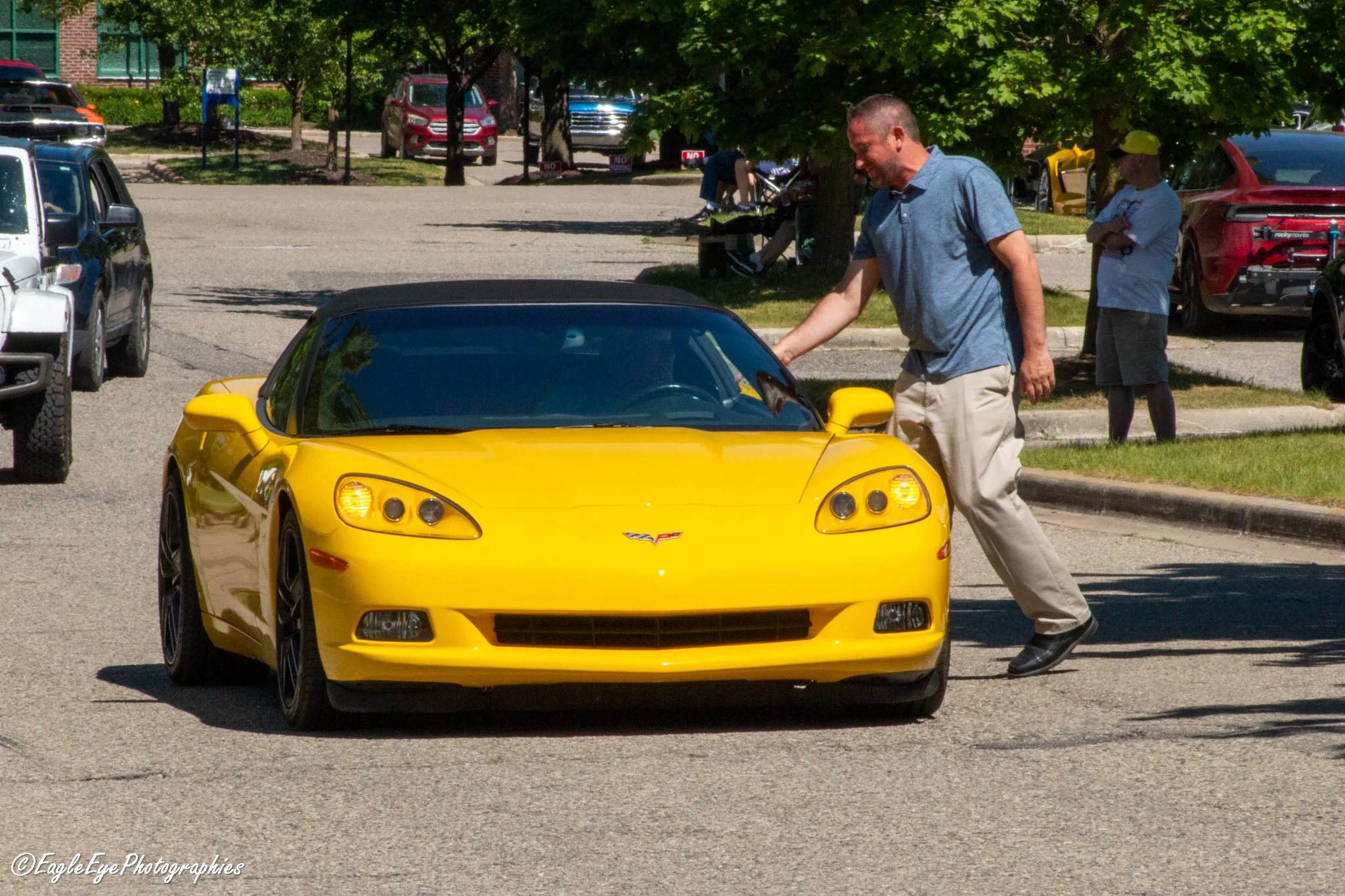 Man handing a yellow sports car to a woman in a parking lot under a tree