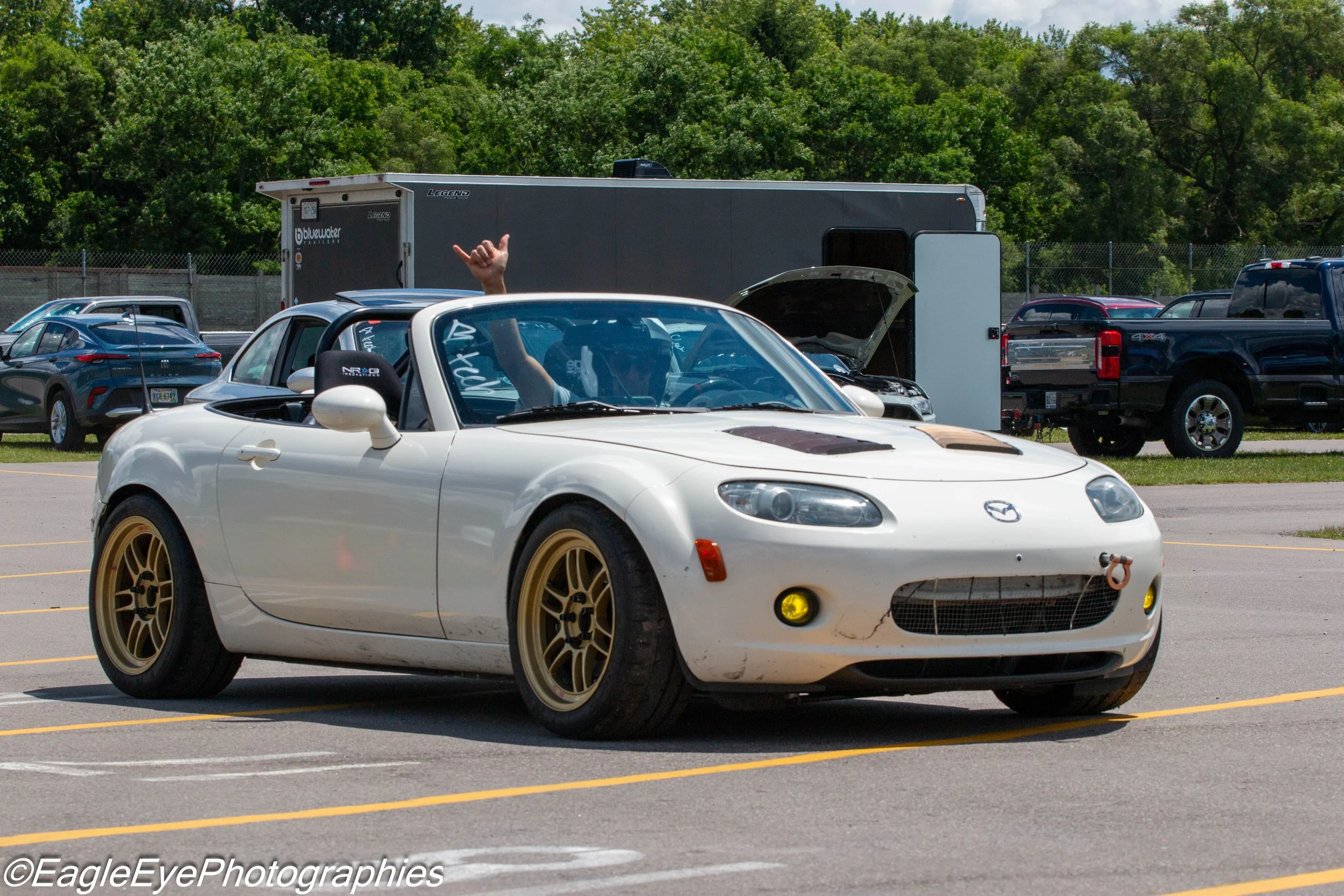 A white Mazda MX-5 Miata convertible sports car with gold rims parked in a parking lot, with a person inside making a hand gesture through the window, surrounded by several other cars and a large trailer in the background.