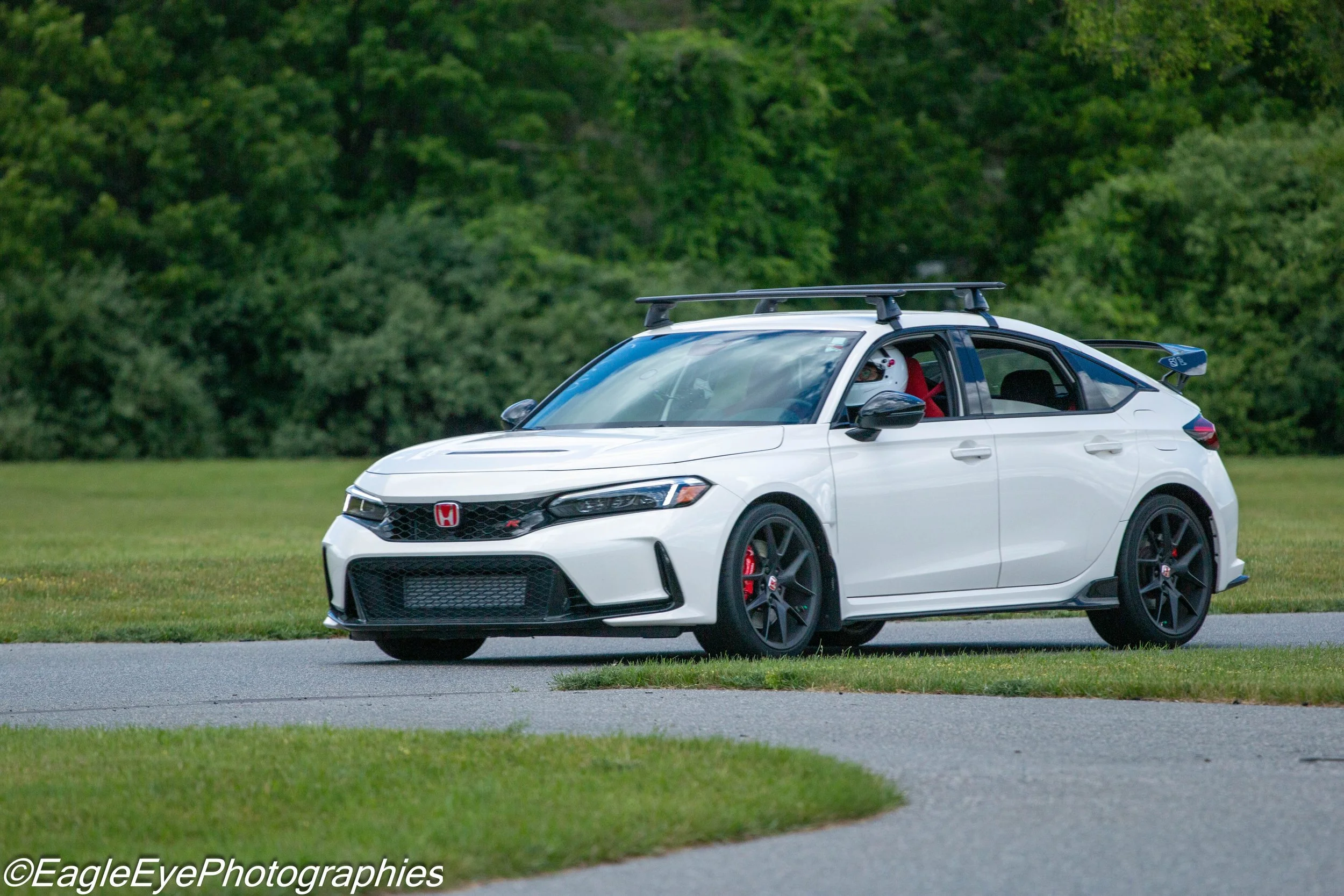 A white sports car with a black front grille and red Honda emblem parked on a paved road beside a grassy area with green trees in the background. The driver is wearing a white helmet and red shirt.