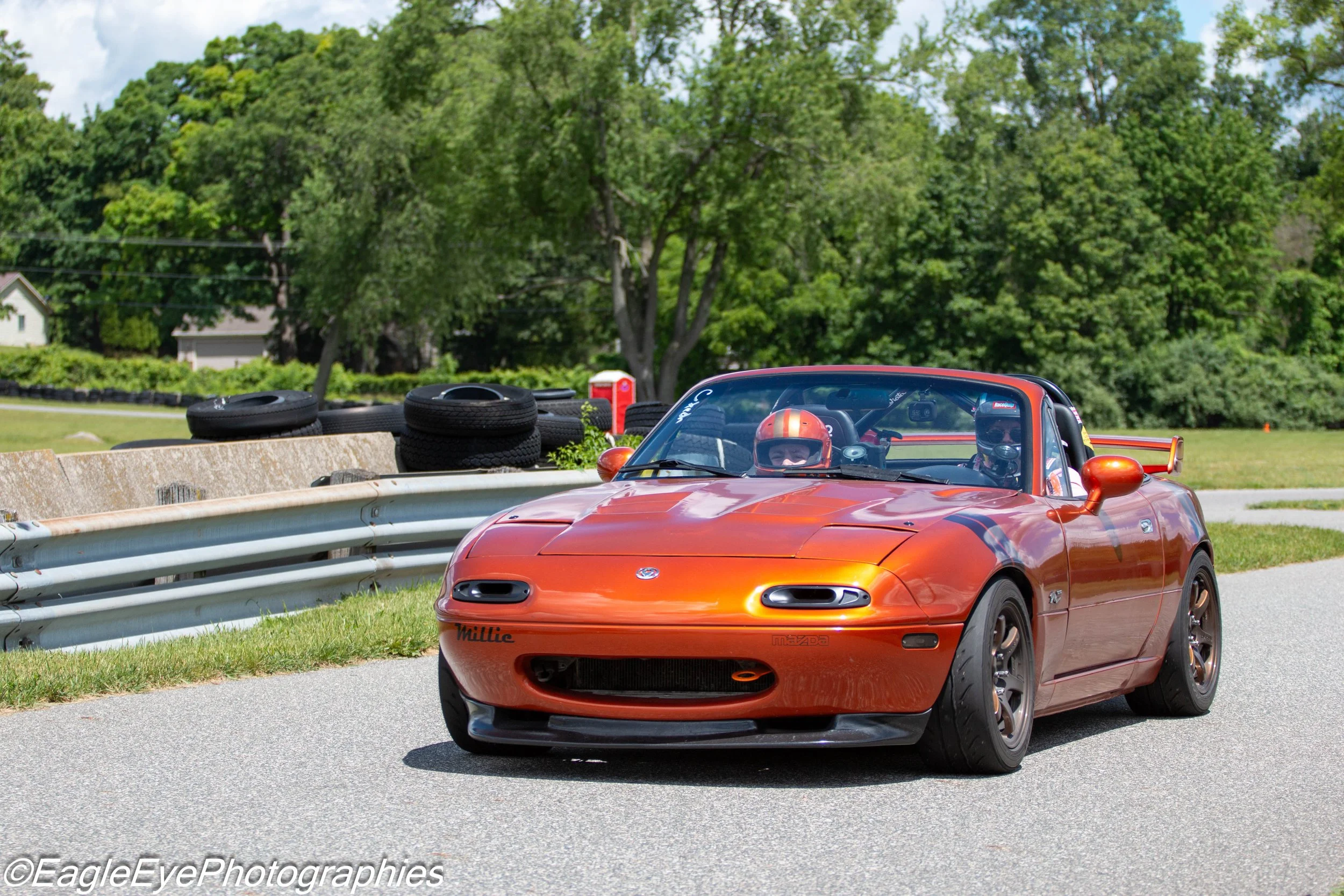 A red Mazda Miata convertible sports car with a driver wearing a helmet is on a race track. The track has a concrete barrier and a stack of tires. In the background, there are lush green trees and a partly cloudy sky.