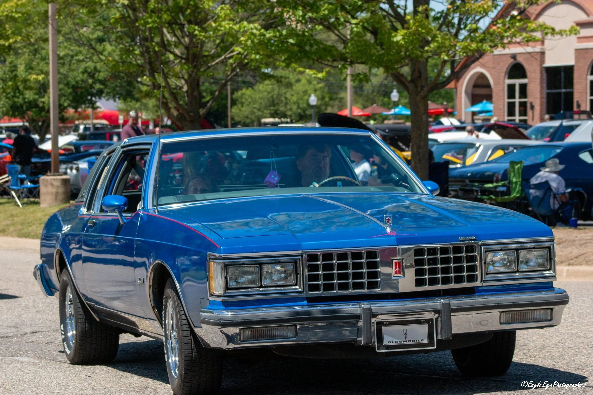 A vintage blue car parked at a car show outdoors with other cars and people in the background, trees, and a building.