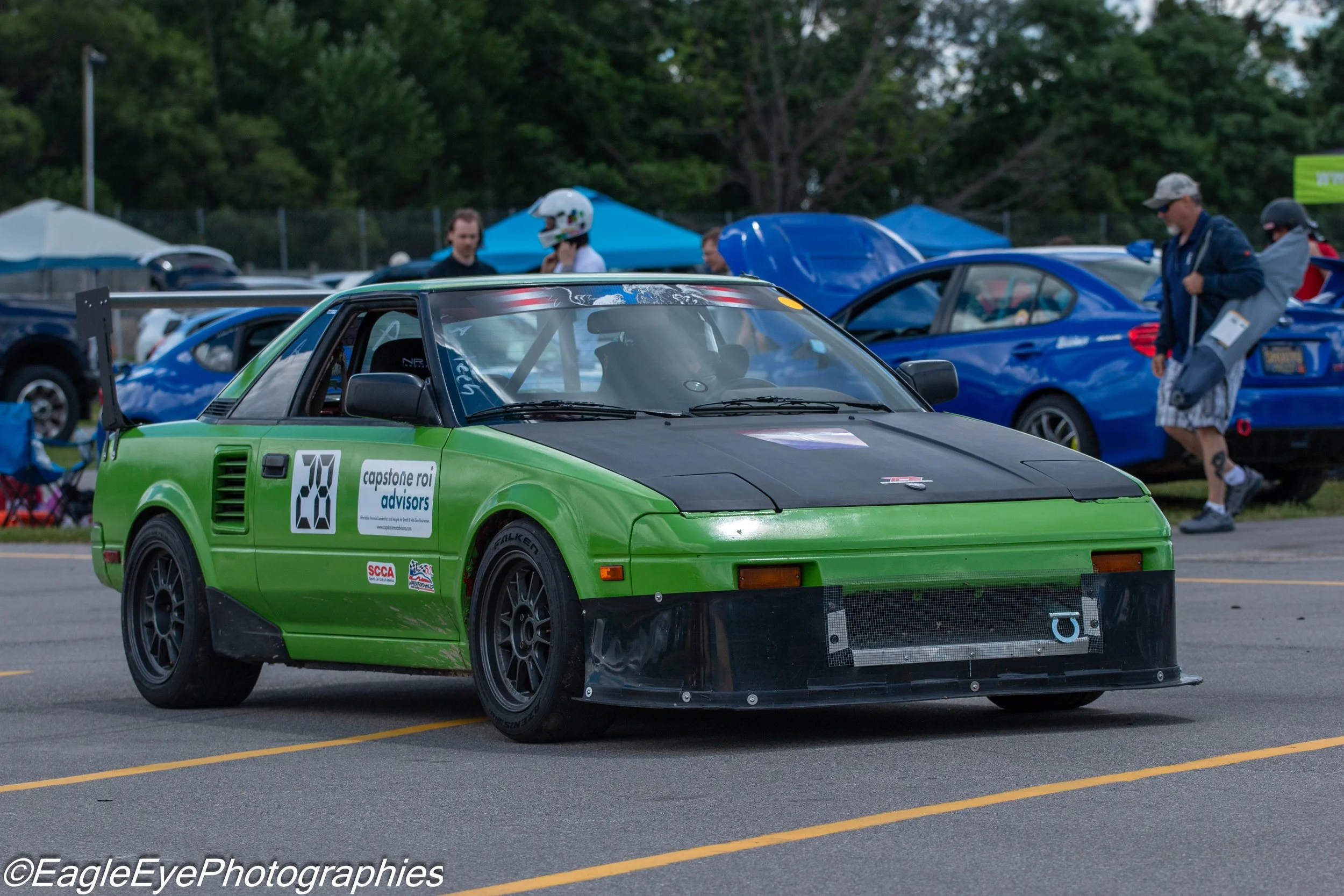 A green race car with the number 28 on its side, parked at a car event with other vehicles and people in the background.