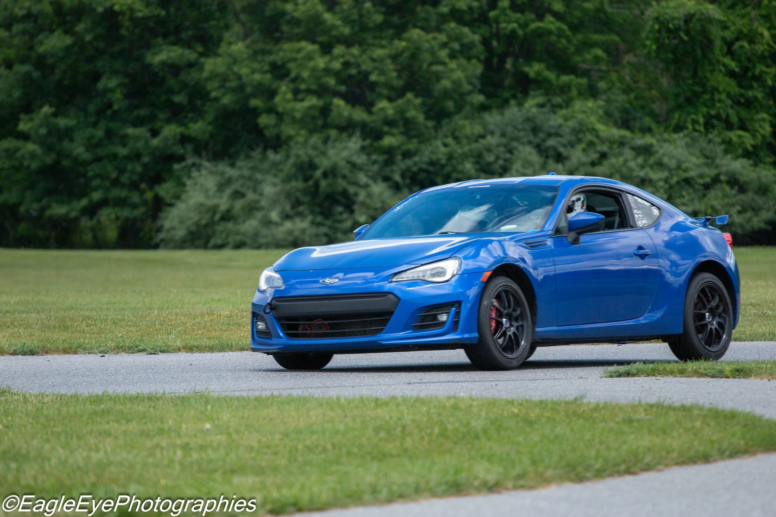A blue sports car driving on a road next to a grassy area with trees in the background.
