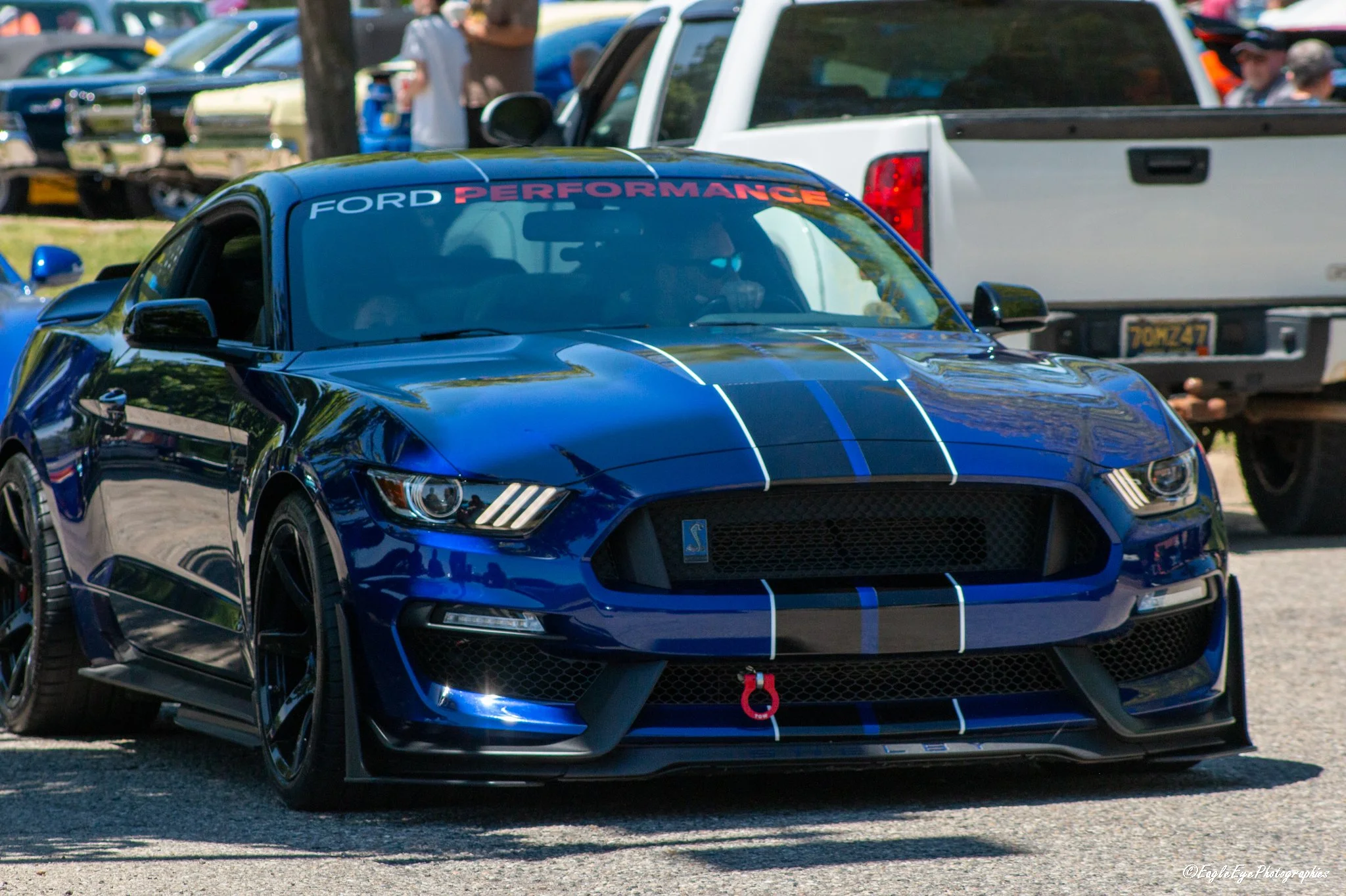 A blue Ford performance sports car with black racing stripes and a red tow hook parked at an outdoor car show, with people and other vehicles in the background.