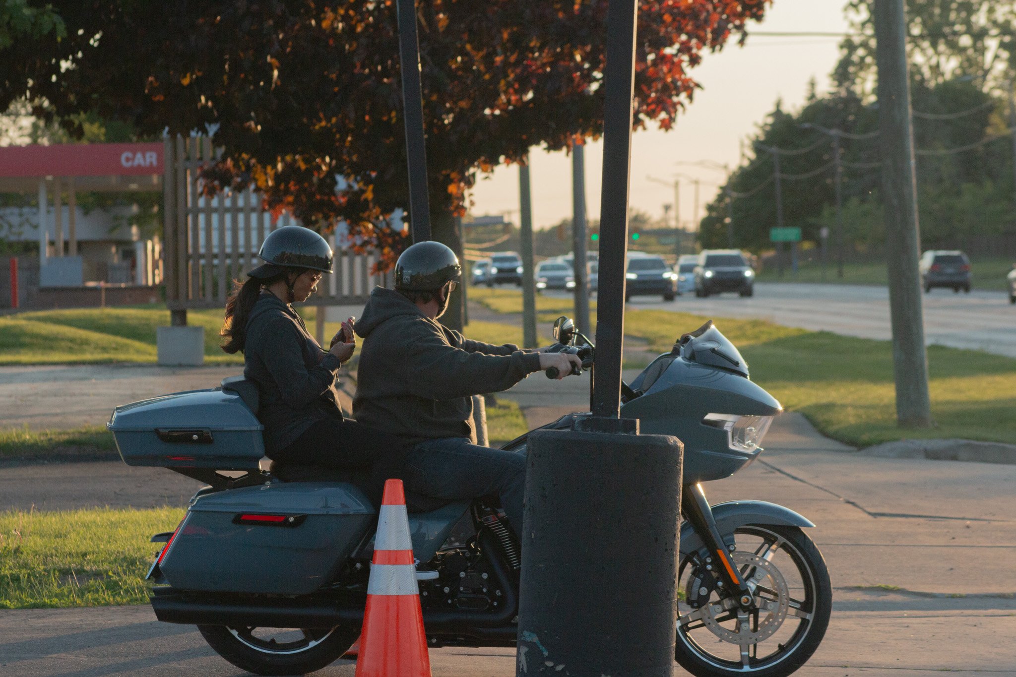 Two people wearing helmets riding a motorcycle past traffic cones on a street during sunset, with cars in the background.