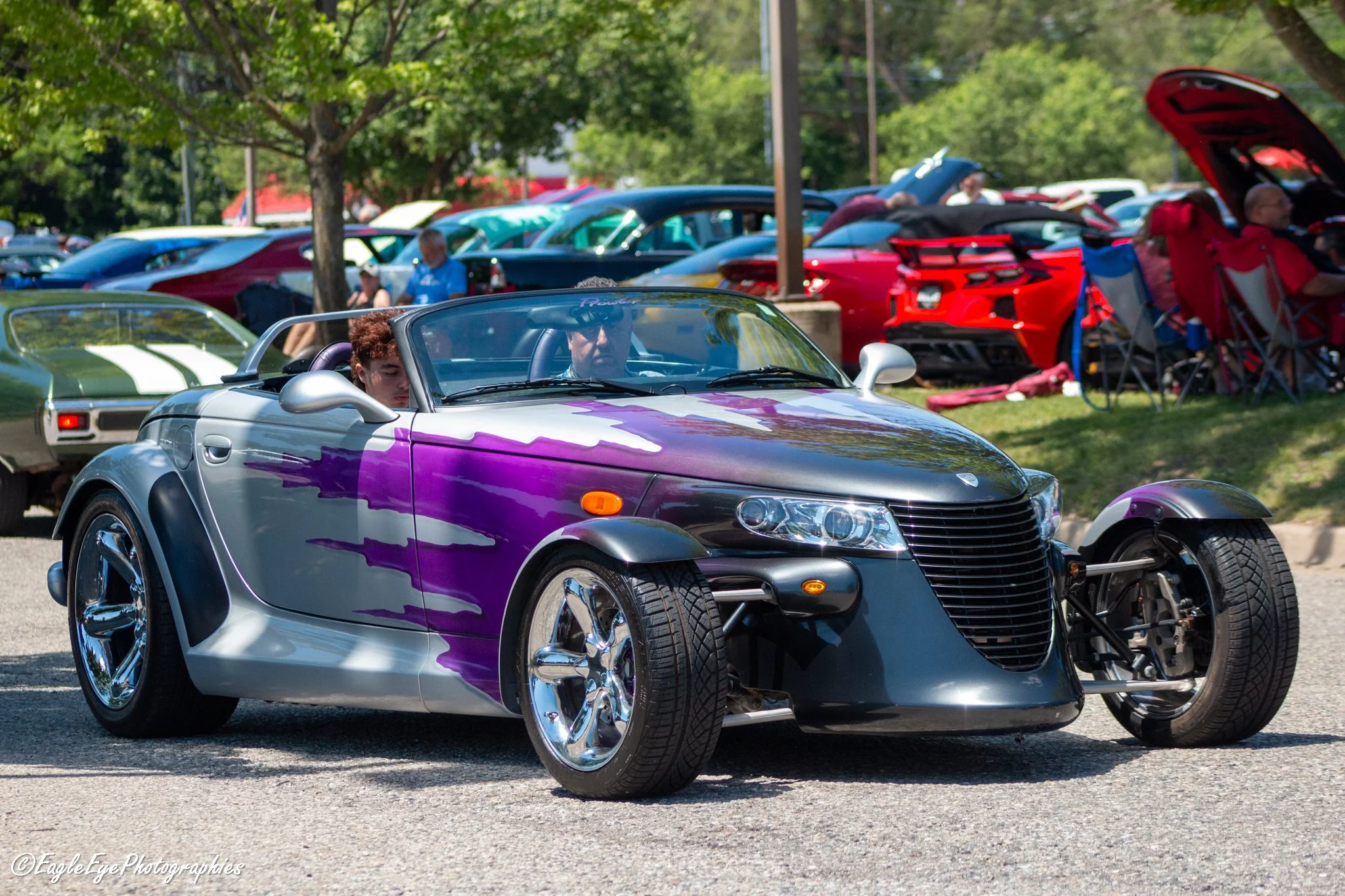 A gray and purple convertible sports car parked at a car show, with people and other classic and sports cars visible in the background under trees.
