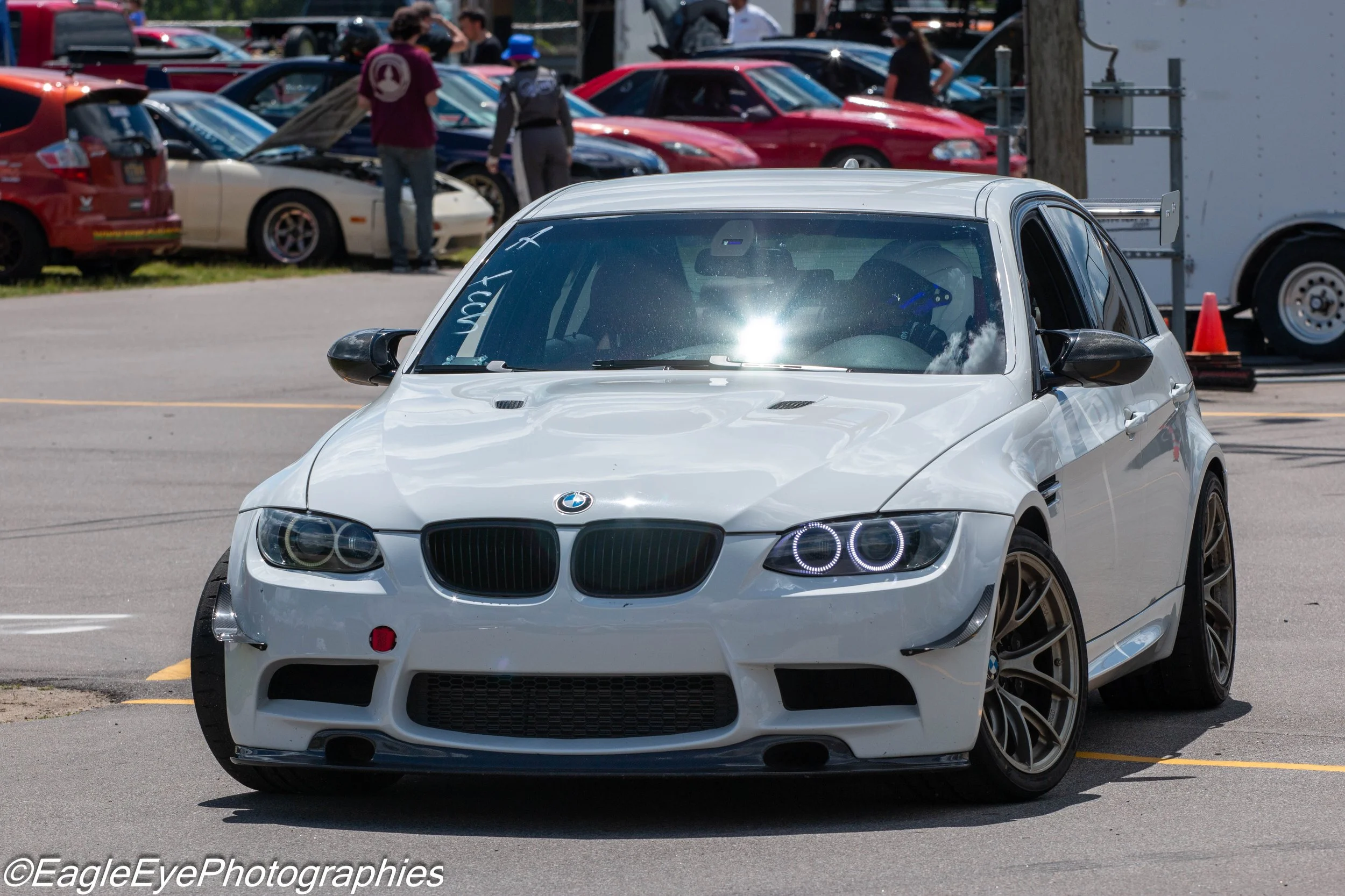 White modified BMW sports car with black accents and halo headlights parked in a lot at a vehicle event. In the background, several other cars and people are visible, some inspecting or talking nearby.