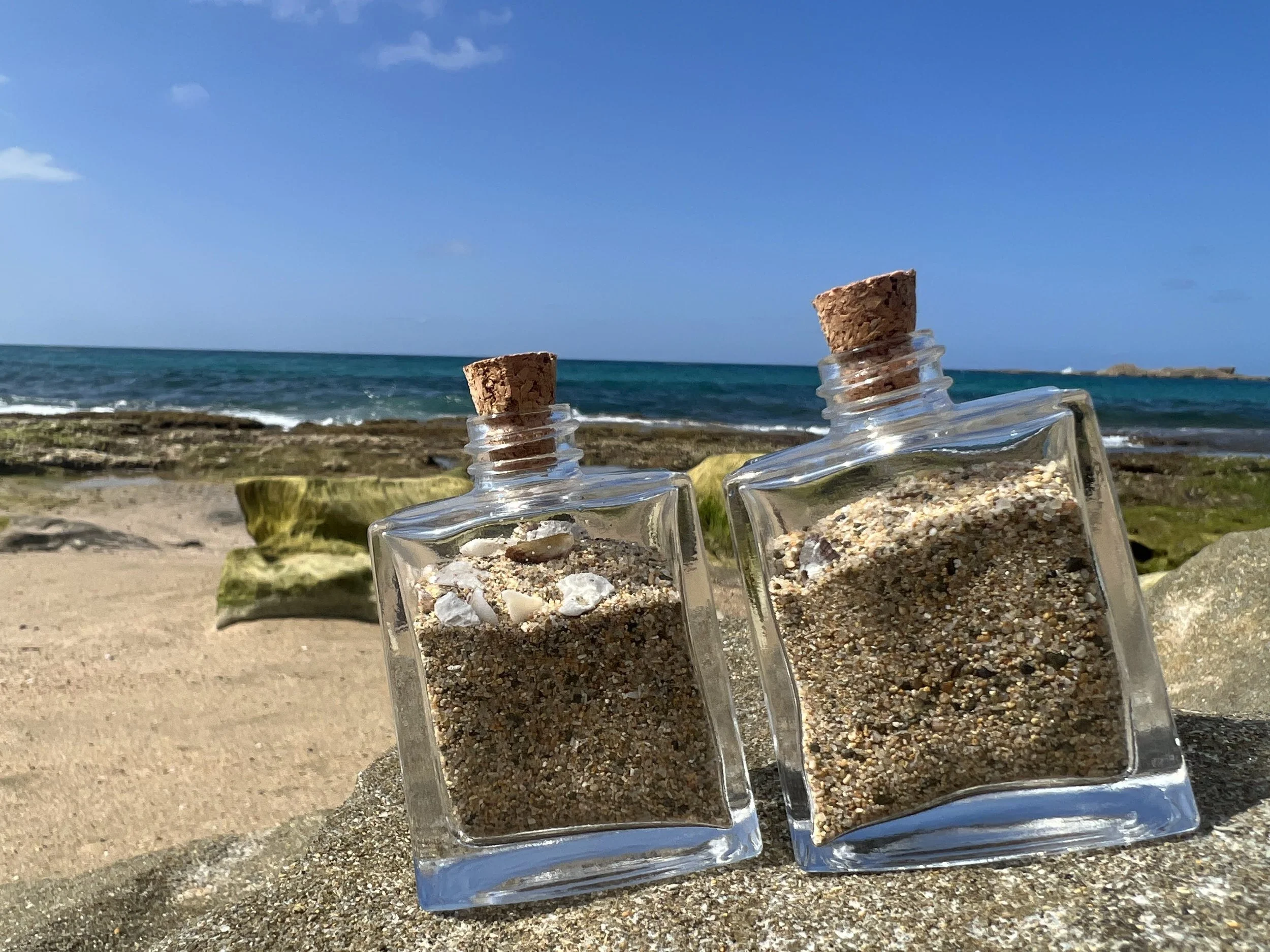 Three small glass bottles filled with sand and small shells, each sealed with a cork, are placed on a beach with the ocean and blue sky in the background.