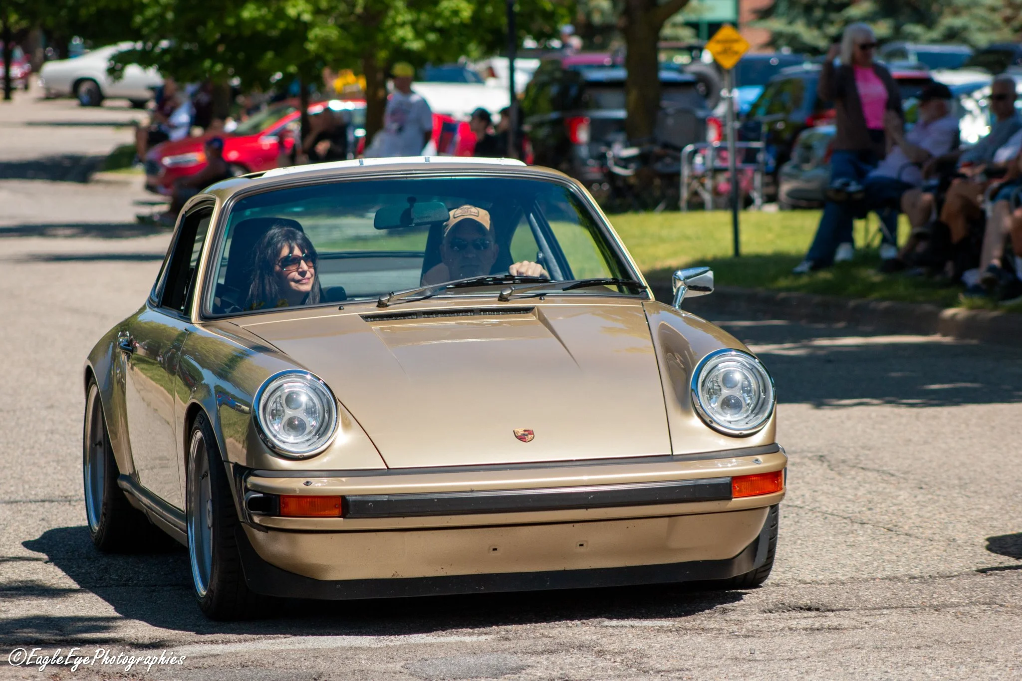 A beige vintage Porsche 911 driving down a street on a sunny day with a crowd of people sitting on benches and standing under trees in the background.