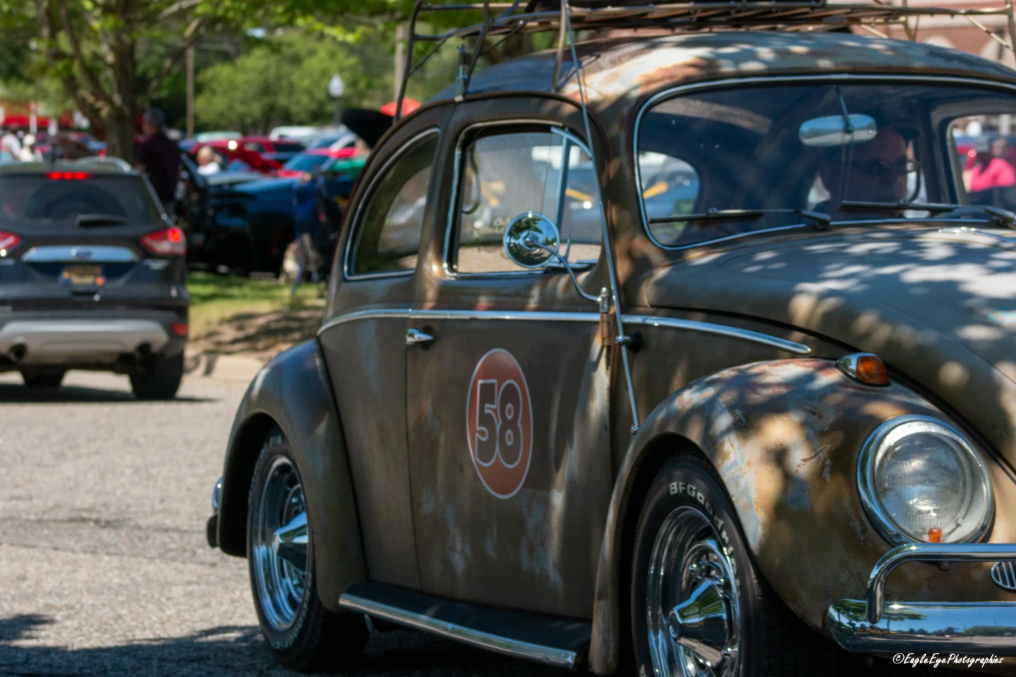 Close-up of a vintage black car with the number 58 on its side, parked among modern vehicles on a street with trees and people in the background.
