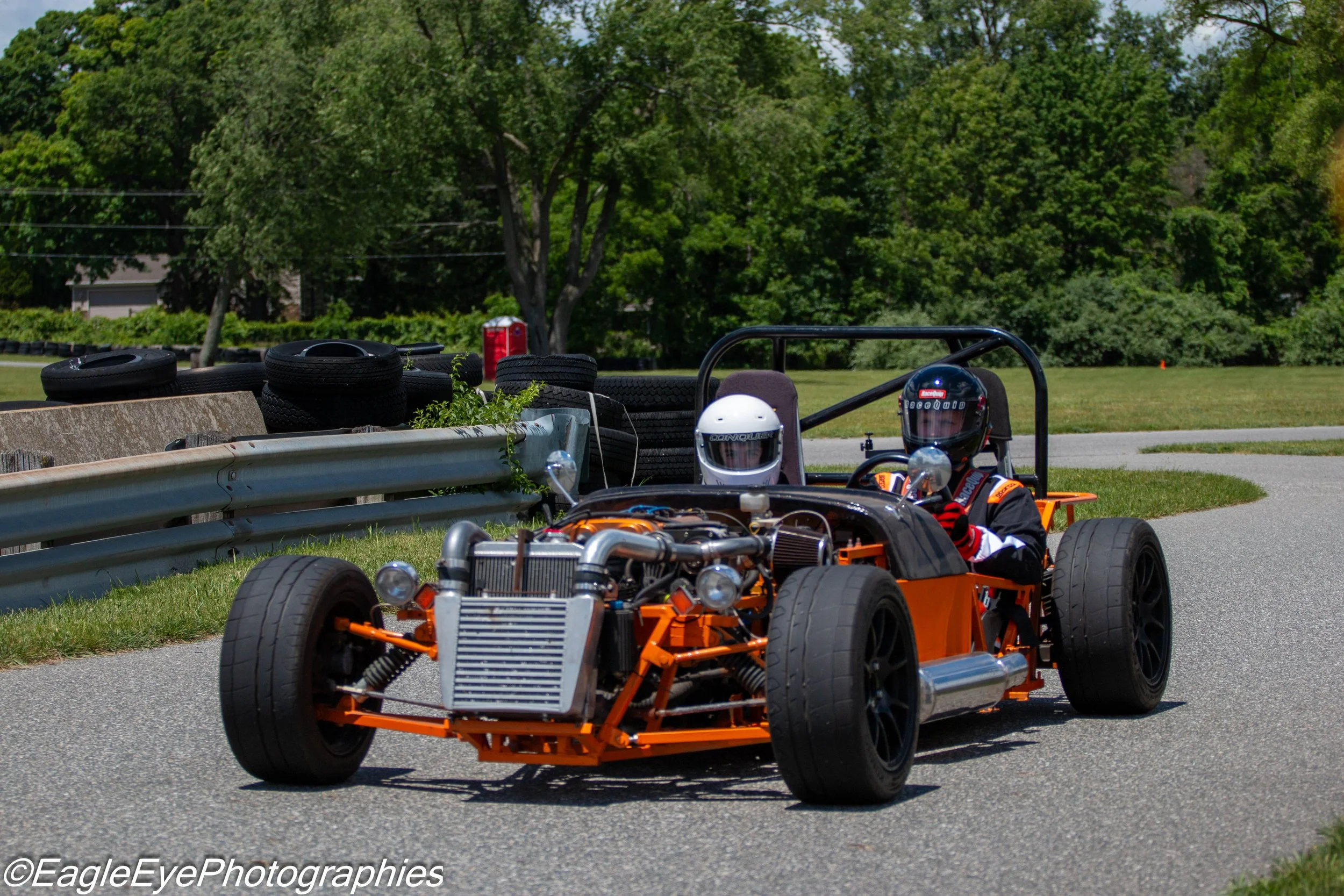 A go-kart race with two children wearing helmets, driving on a paved track. Tire stacks and greenery in the background.