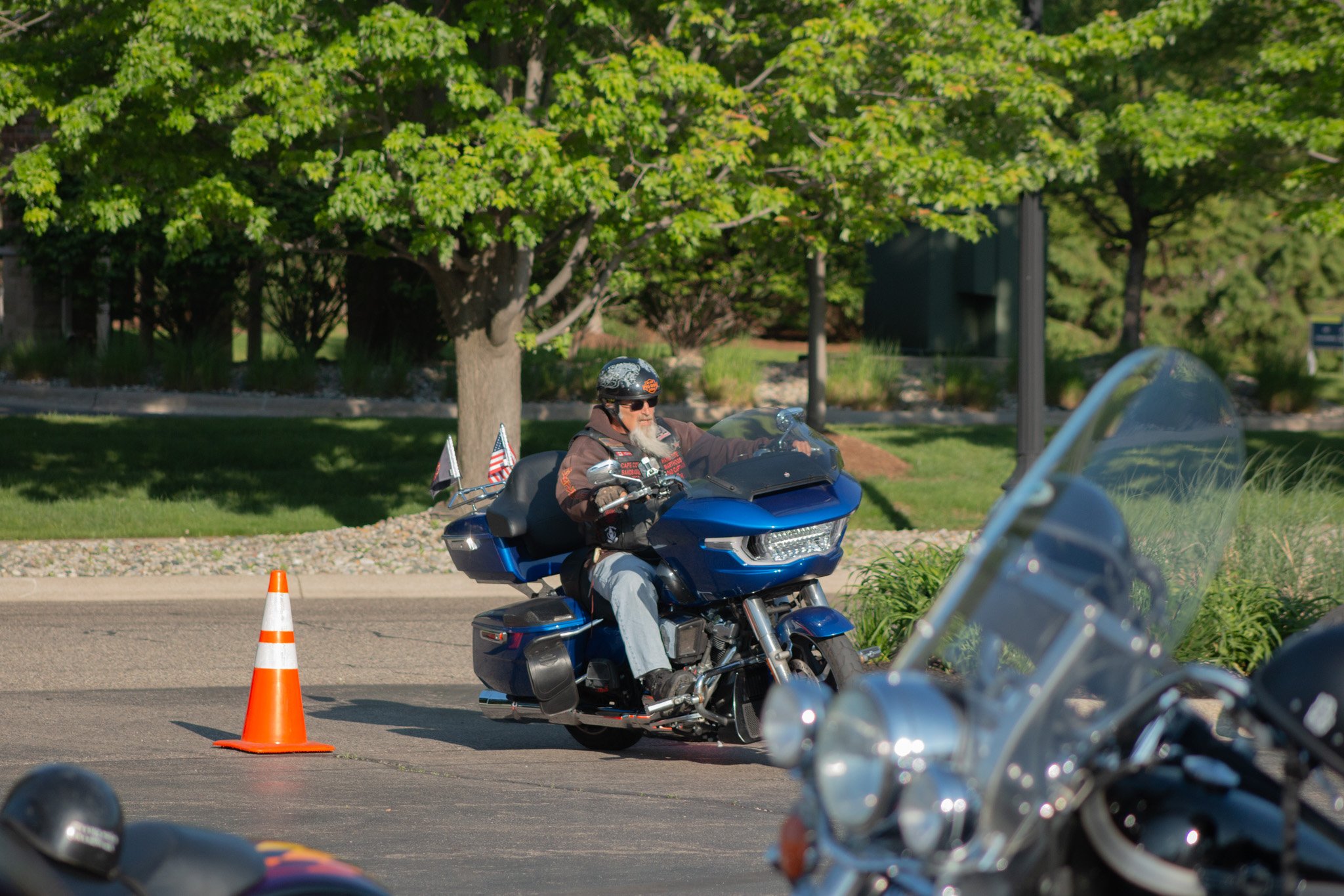 A man with a long white beard wearing sunglasses and a brown jacket riding a blue motorcycle on a street, with orange traffic cones marking the area. Green trees are visible in the background.