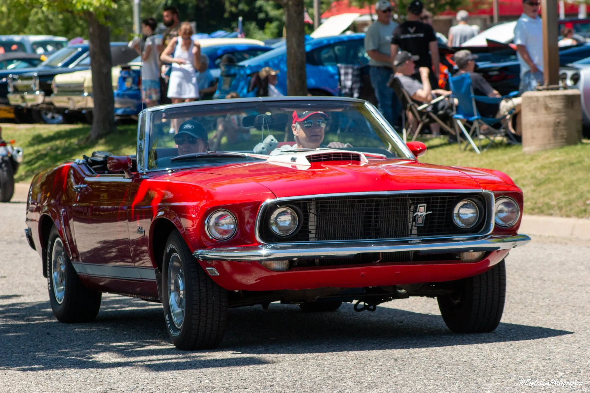 Front view of a red vintage convertible car with a man and a woman inside, participating in a car show with a crowd in the background.