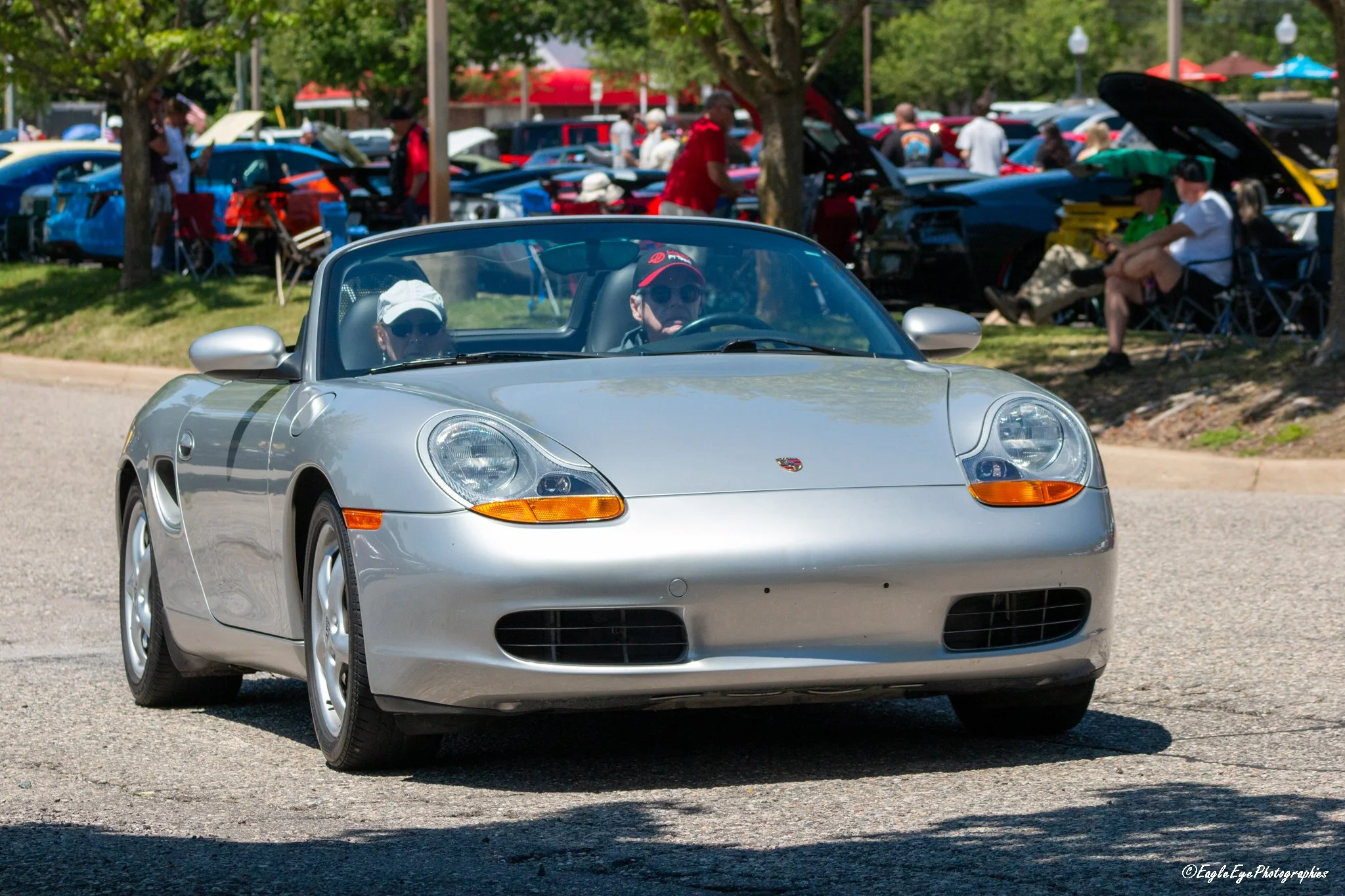 A silver Porsche convertible driving in a car show with people sitting and walking in the background, some with open car hoods and tents, under trees on a sunny day.