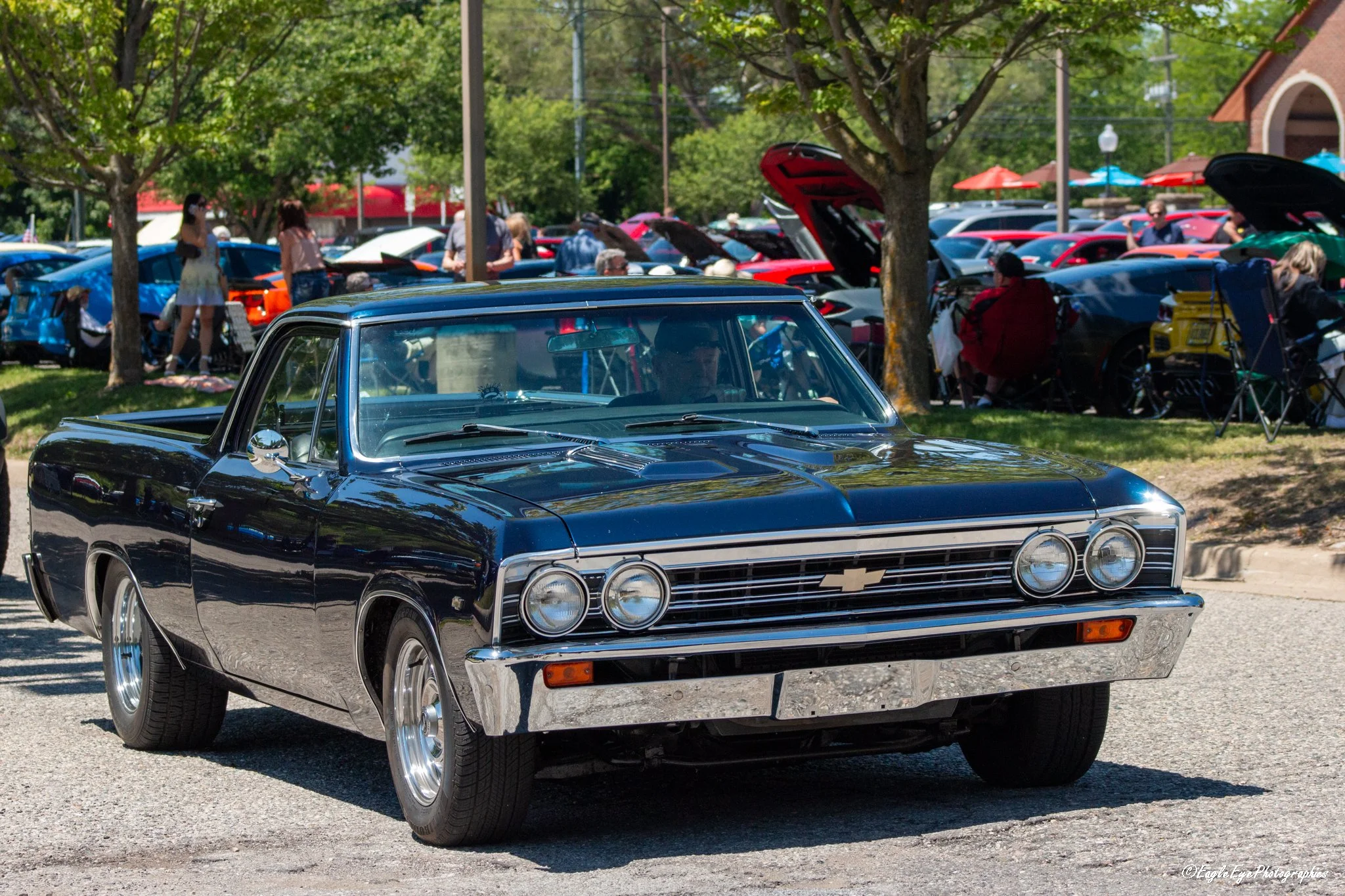 A black vintage Chevrolet car parked in the foreground at a car show, with people and more cars, some with open hoods, and trees in the background on a sunny day.