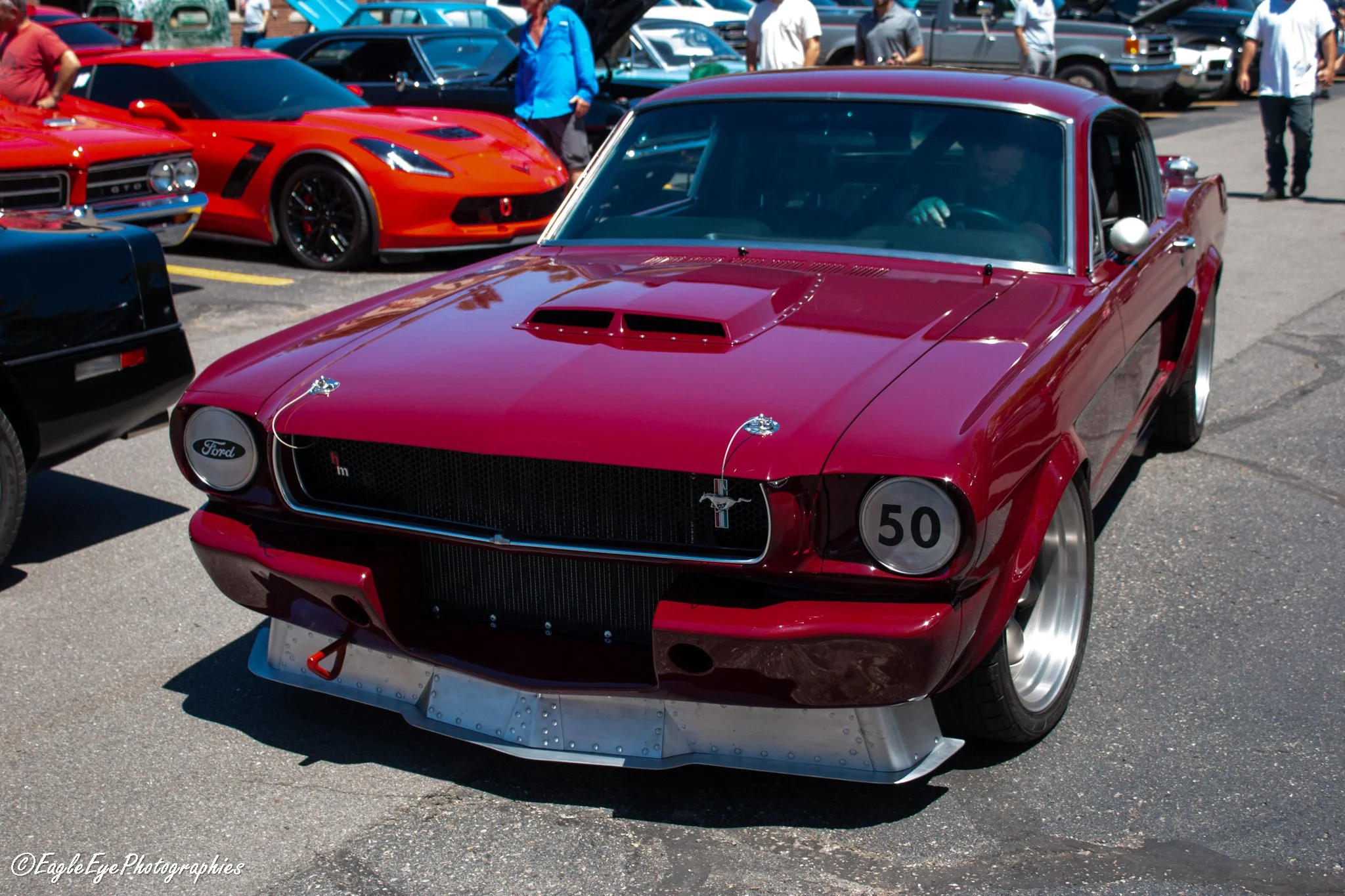A red classic race car with a vented hood and metallic fins on the front, parked among other vintage vehicles at a car show, with people in casual clothing walking in the background.