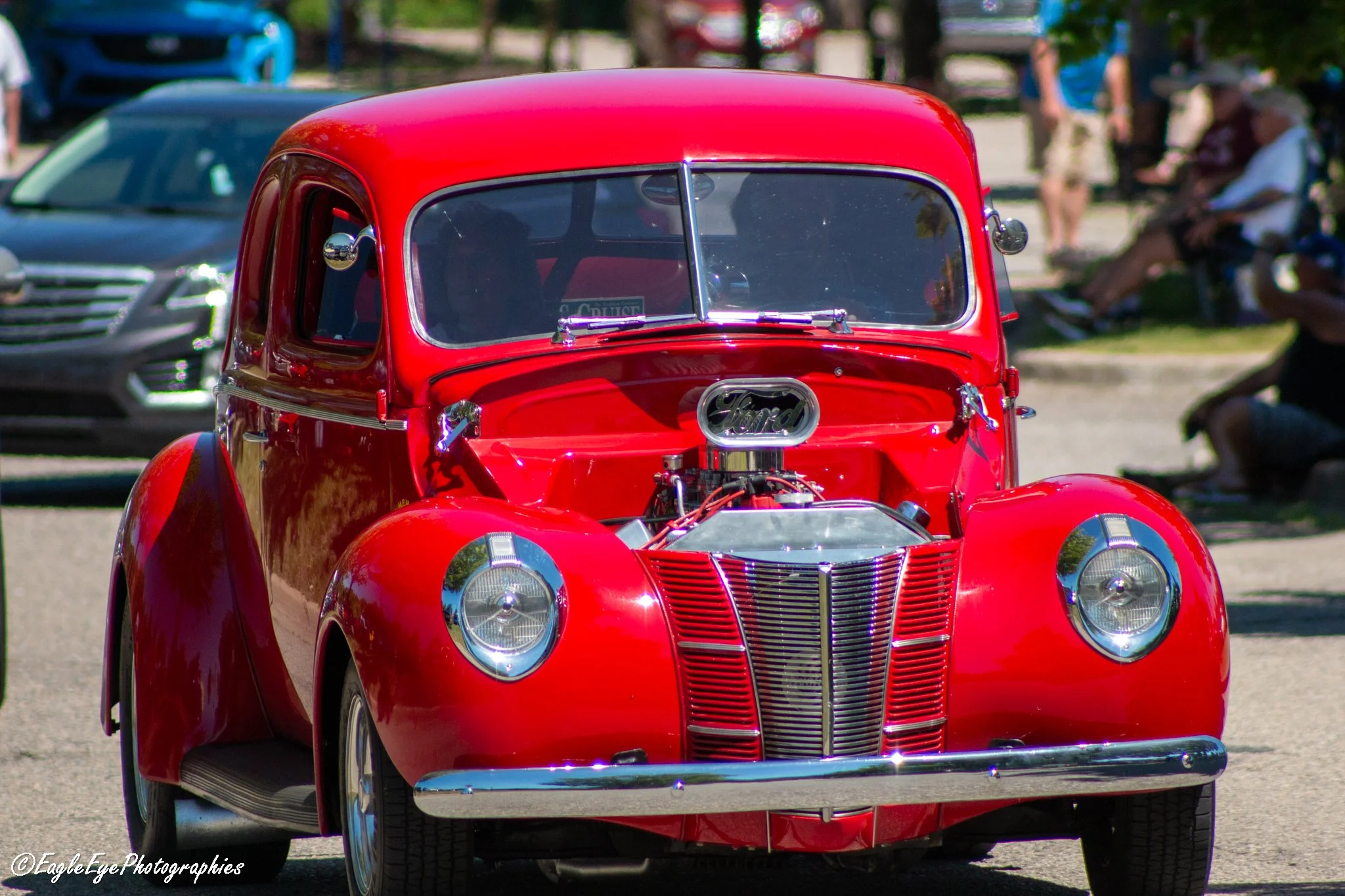 A close-up of a vintage red Ford car on display at an outdoor event, with other cars and spectators in the background.