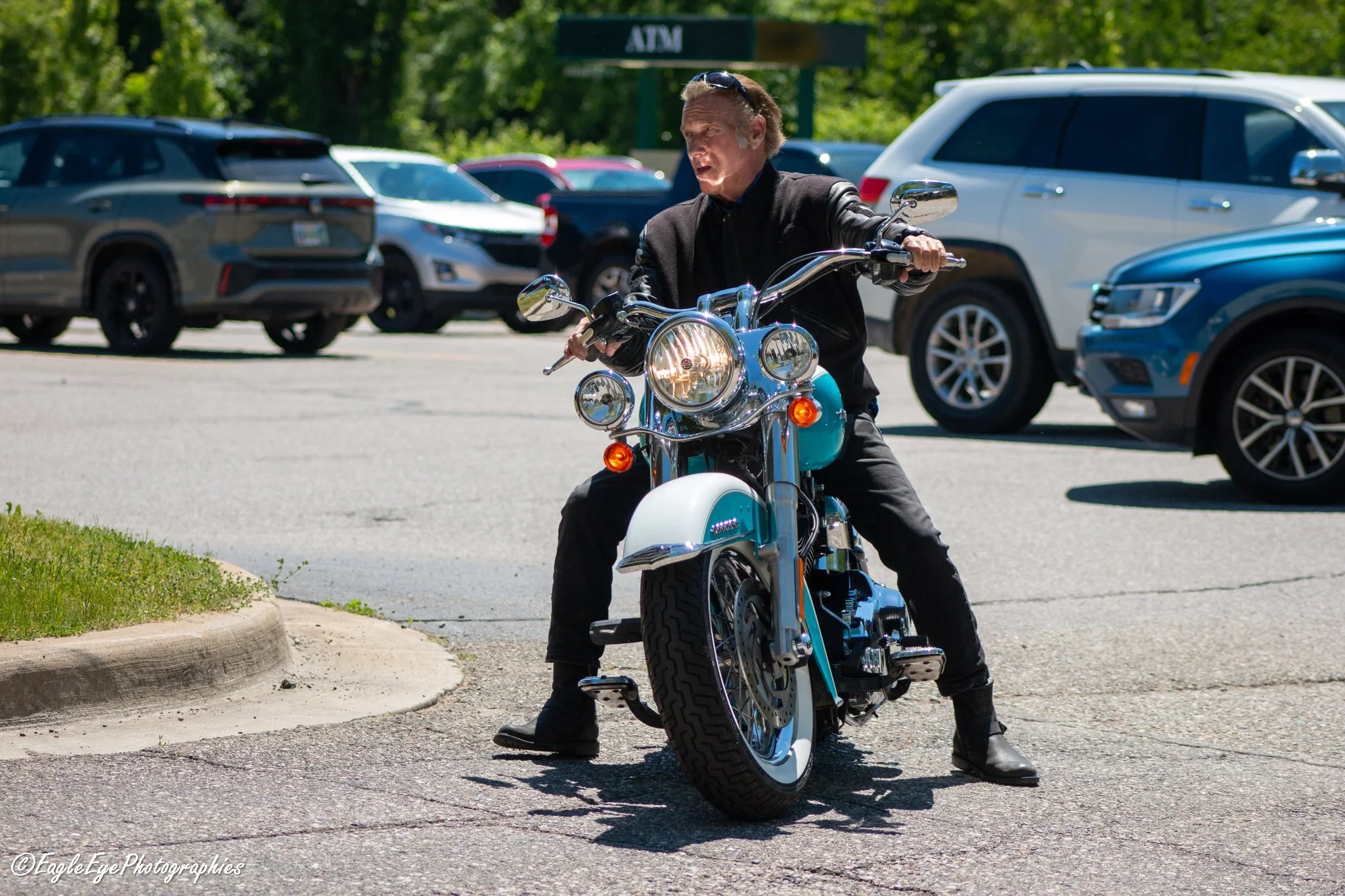 A man riding a turquoise and chrome motorcycle in a parking lot with parked cars and green trees in the background.