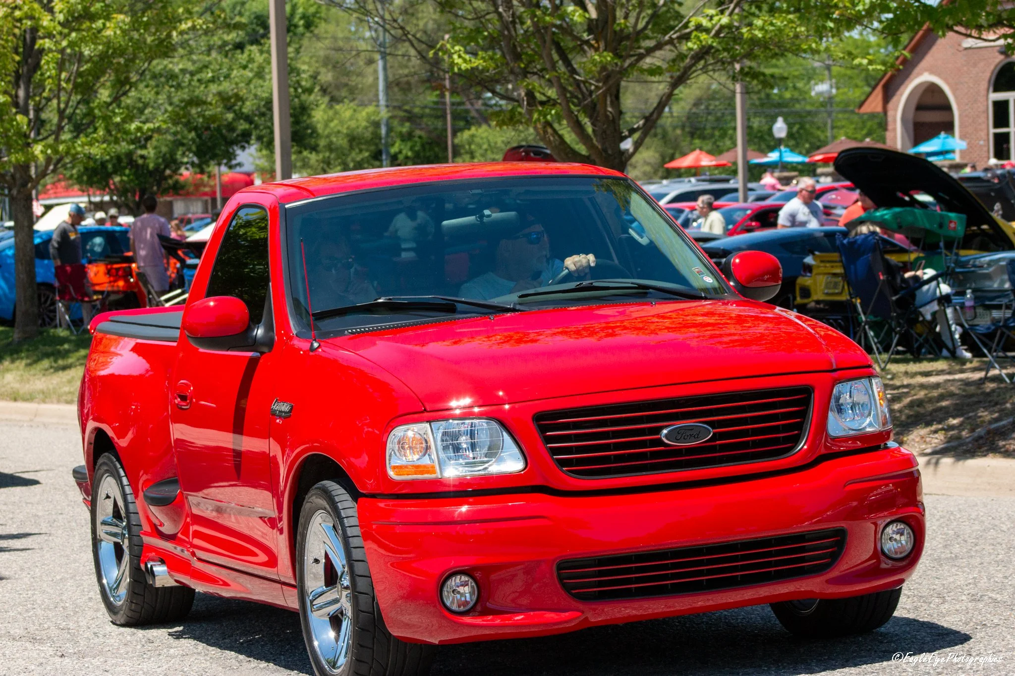 A red Ford pickup truck parked in a crowded outdoor event, with trees and people in the background.