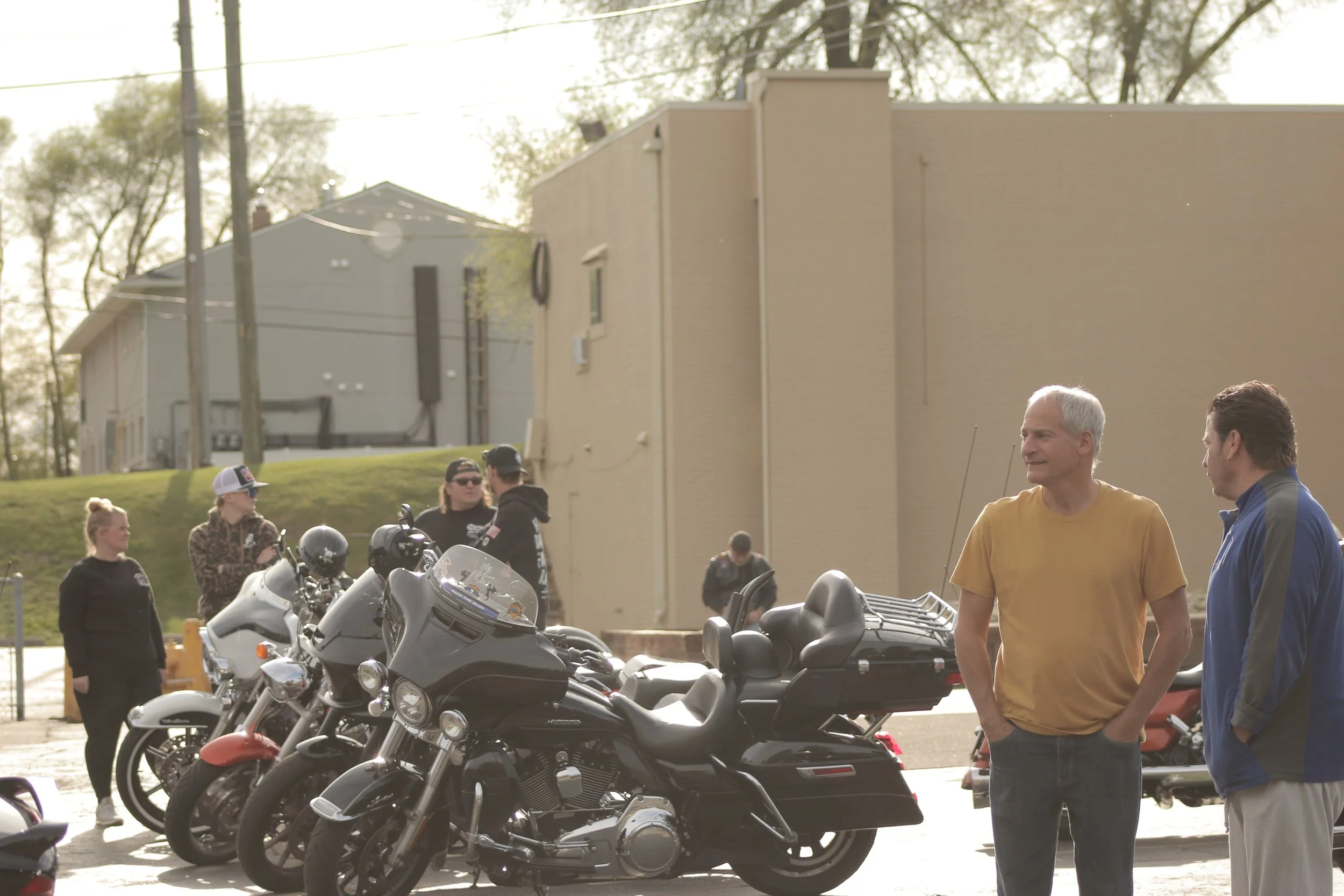 People standing near motorcycles parked in a lot outdoors during daytime, with buildings and trees in the background.