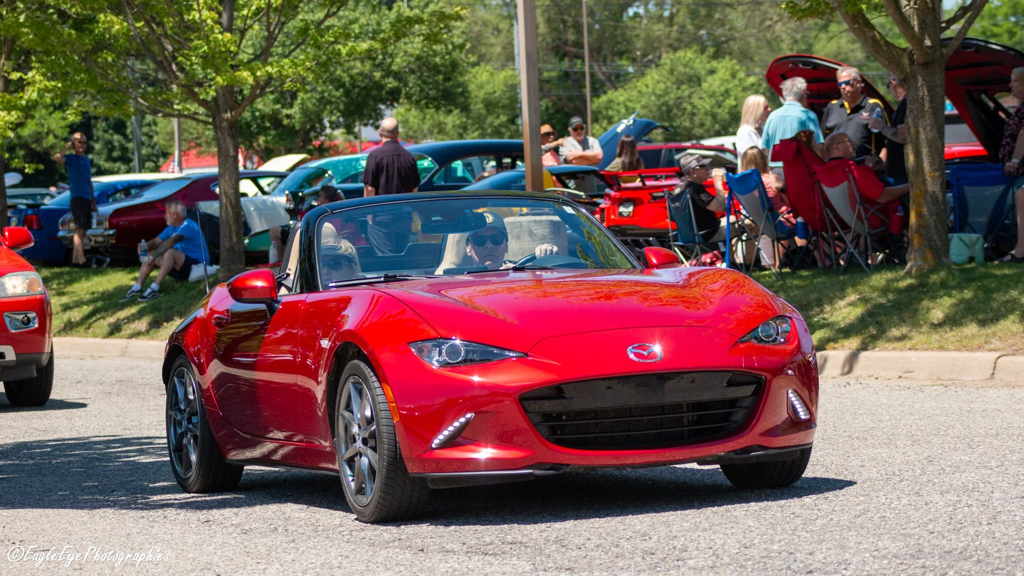 Red convertible Mazda sports car driving past a car show with people gathered around vintage and modern cars on a grassy area under trees on a sunny day.
