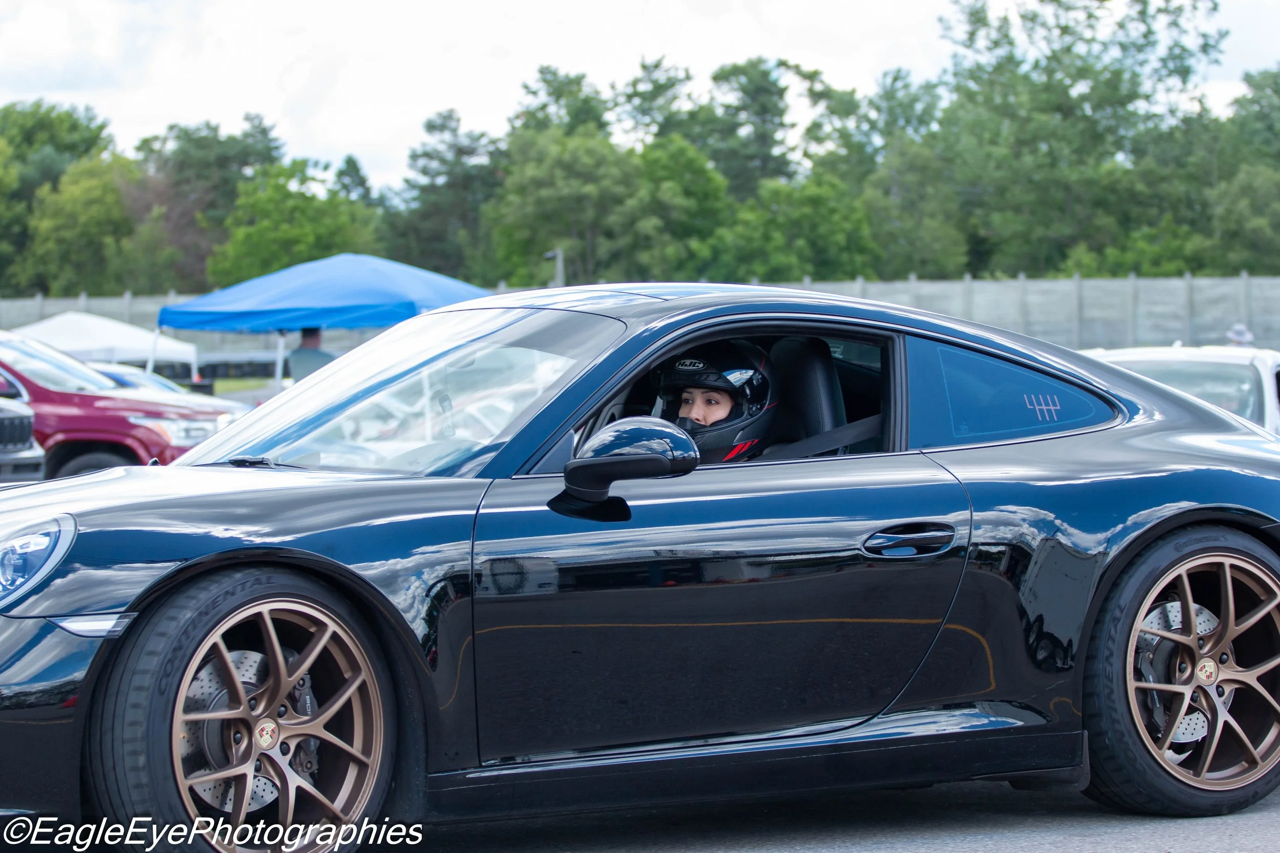 A person inside a black sports car wearing a helmet at a racing event, with other parked vehicles and trees in the background.