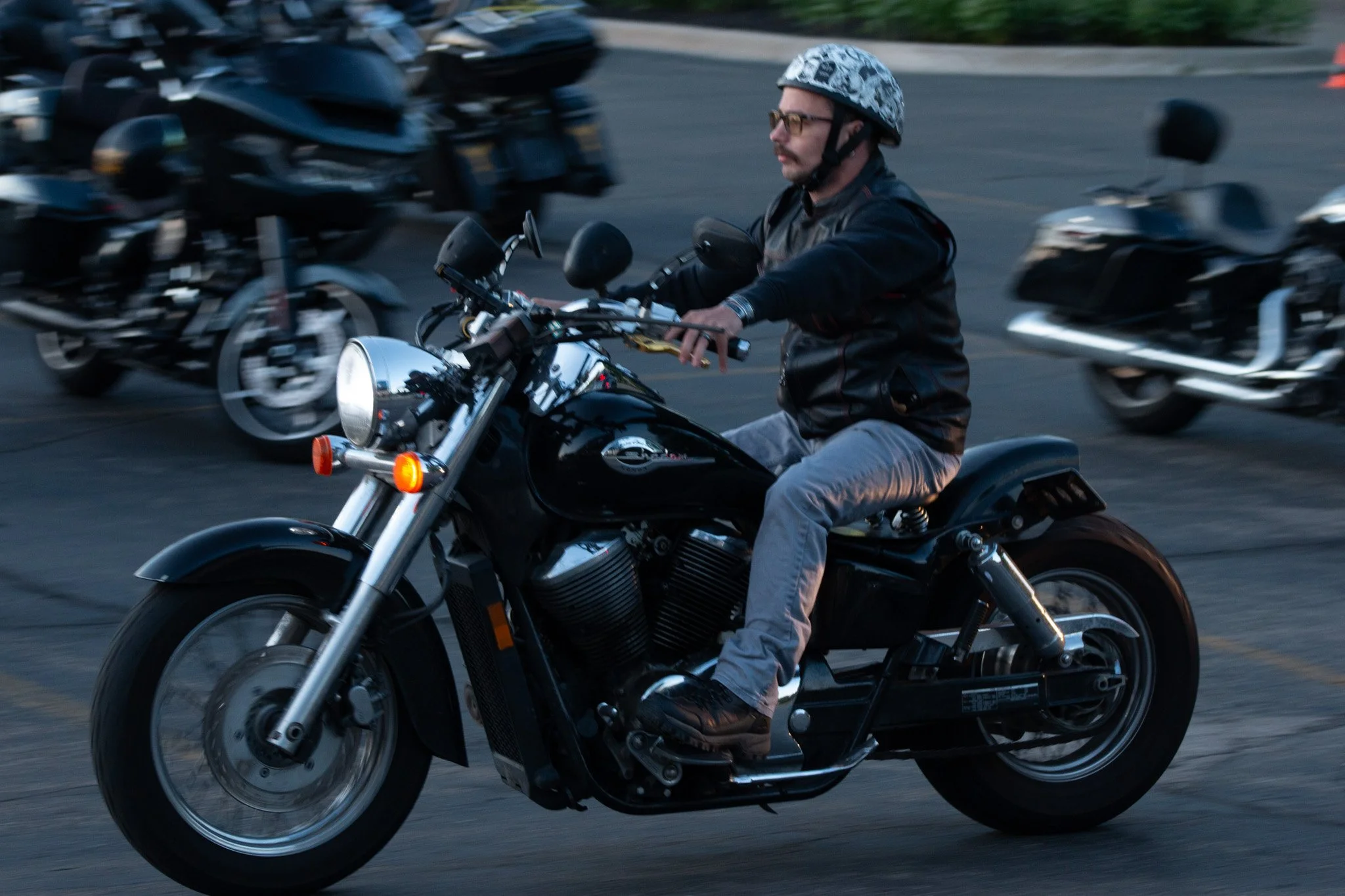 A man riding a black cruiser motorcycle with a white headlight, wearing glasses, a patterned helmet, a black leather jacket, and gray pants, riding in a parking lot with multiple motorcycle parked in the background.