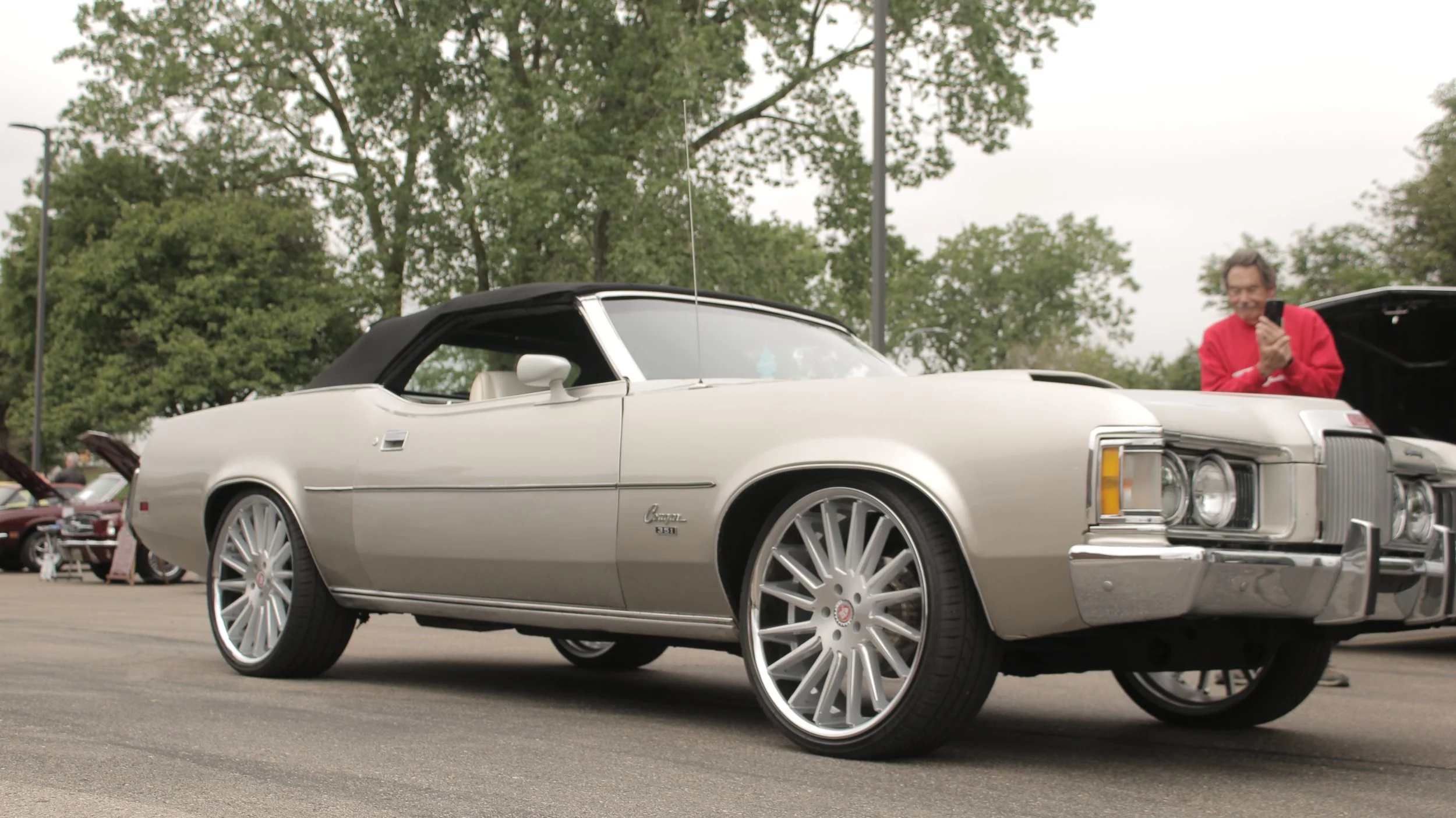 A classic white convertible car with large silver rims parked in a lot during an outdoor event, with a person in a red sweater smiling and taking a photo nearby, trees and other cars in the background.