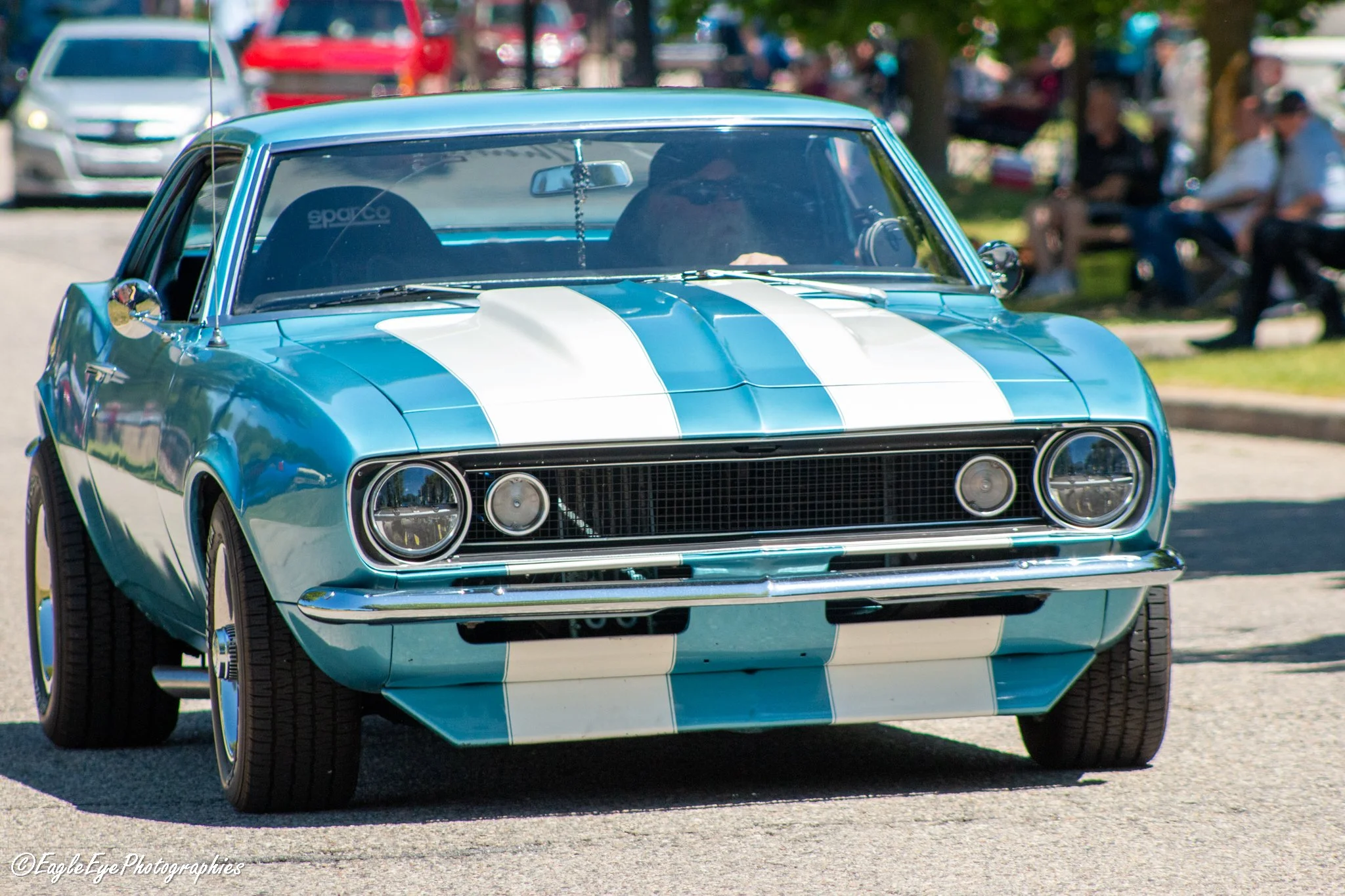 A vintage blue sports car with white racing stripes, parked outdoors with people sitting on benches in the background.