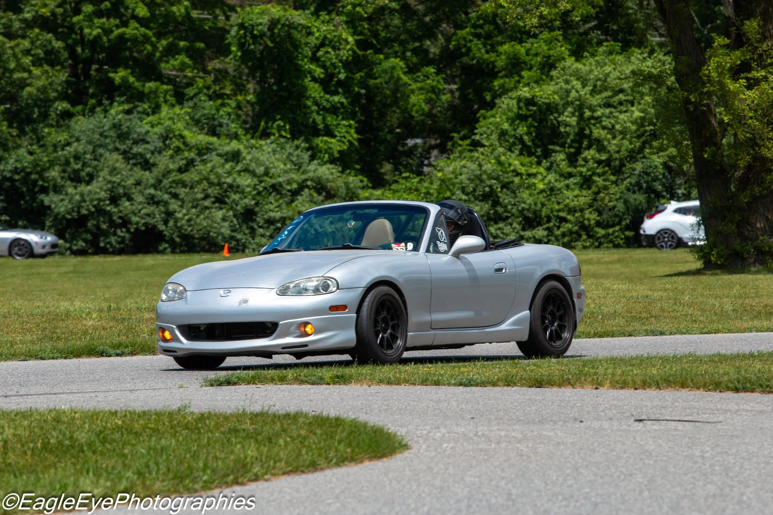 A silver convertible sports car driving on a road with green grass and trees in the background.