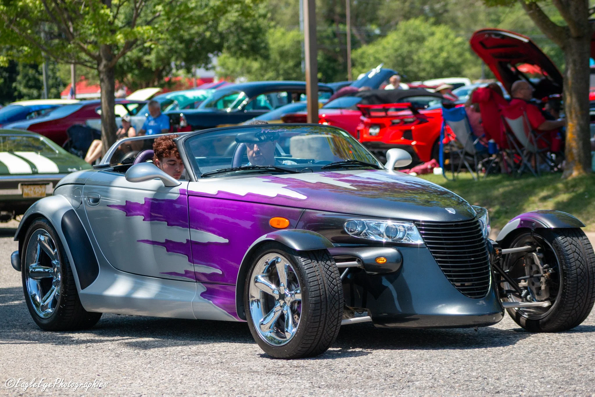 A silver convertible sports car with purple flame decals parked among other classic cars at an outdoor car show on a sunny day, with people and trees in the background.