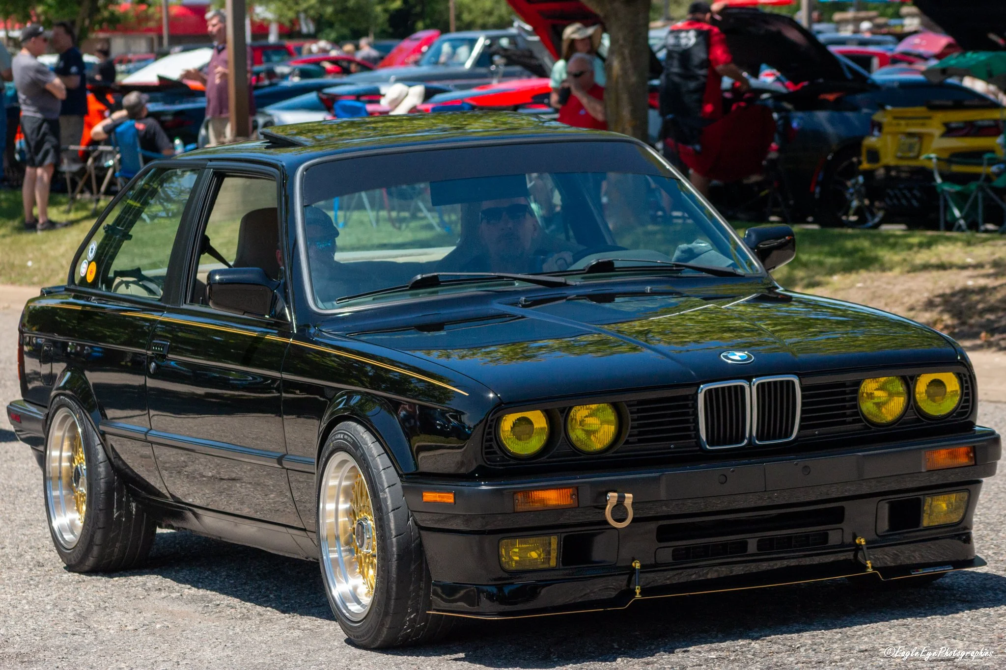 A black vintage BMW car with yellow-tinted headlights and gold rims parked at a car show, with people and other cars visible in the background.