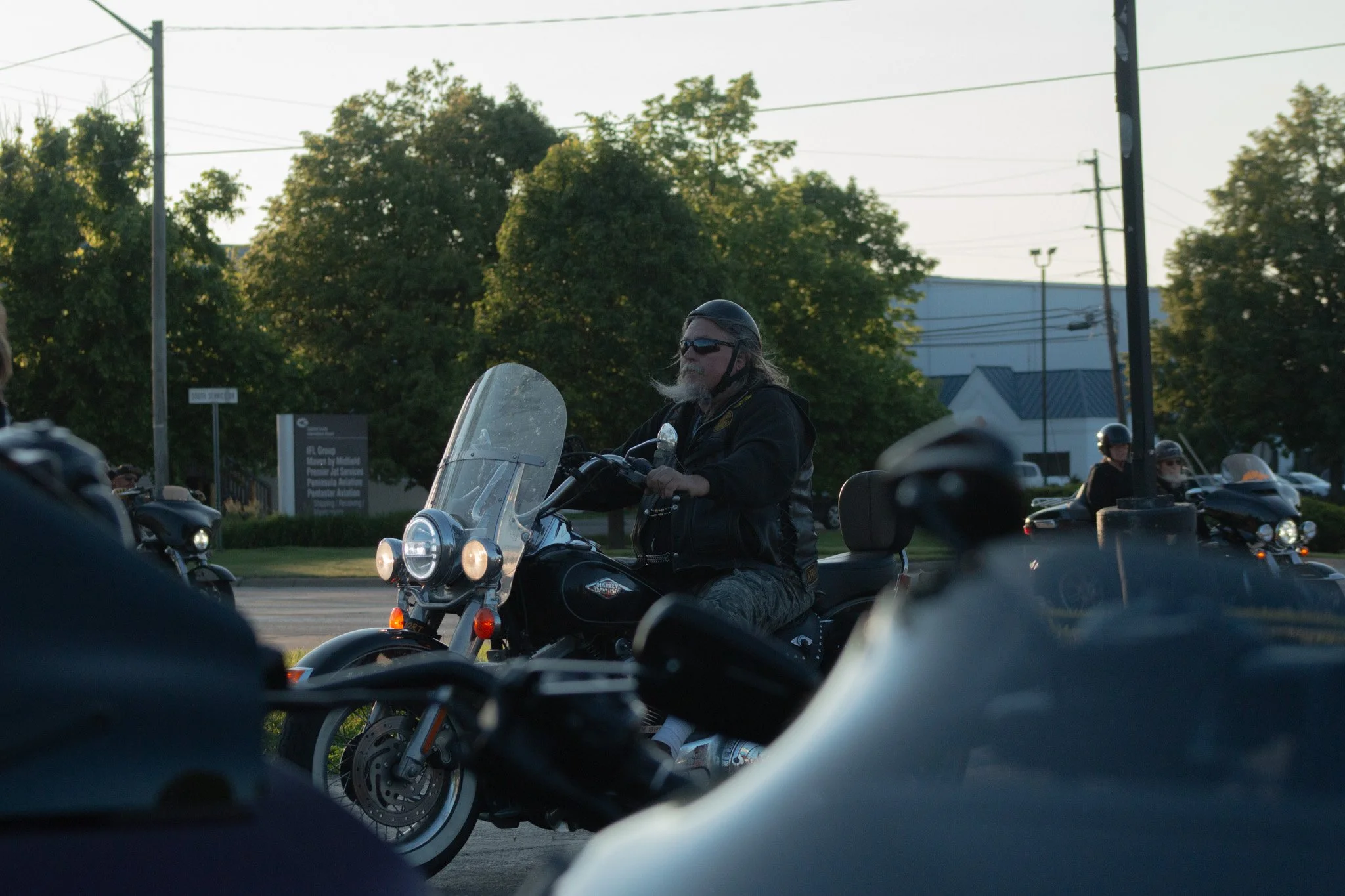 A man with long hair, a beard, sunglasses, and a black helmet riding a motorcycle, with several other motorcycles and riders in the background during dusk or early evening.