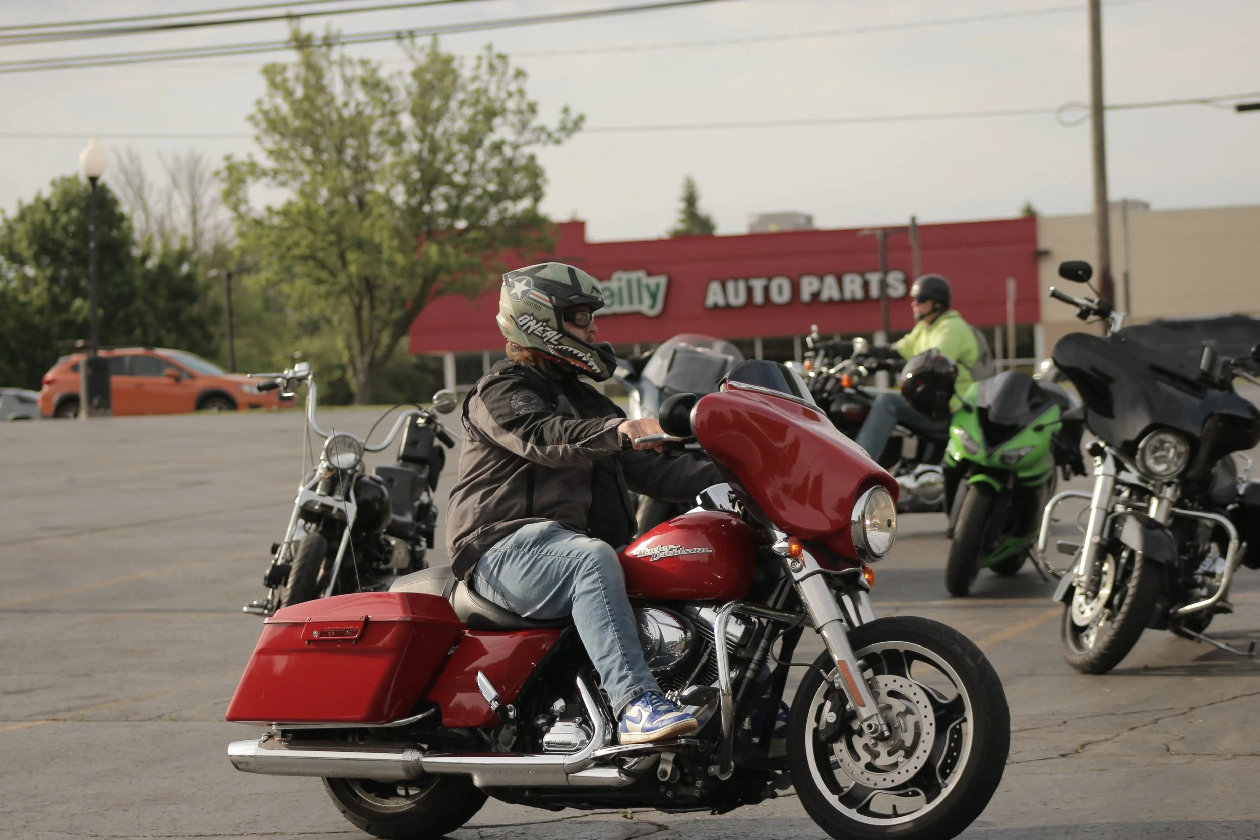 A person wearing a helmet and black riding jacket sitting on a red motorcycle in a parking lot, with other motorcycles and a sign for Auto Parts in the background.