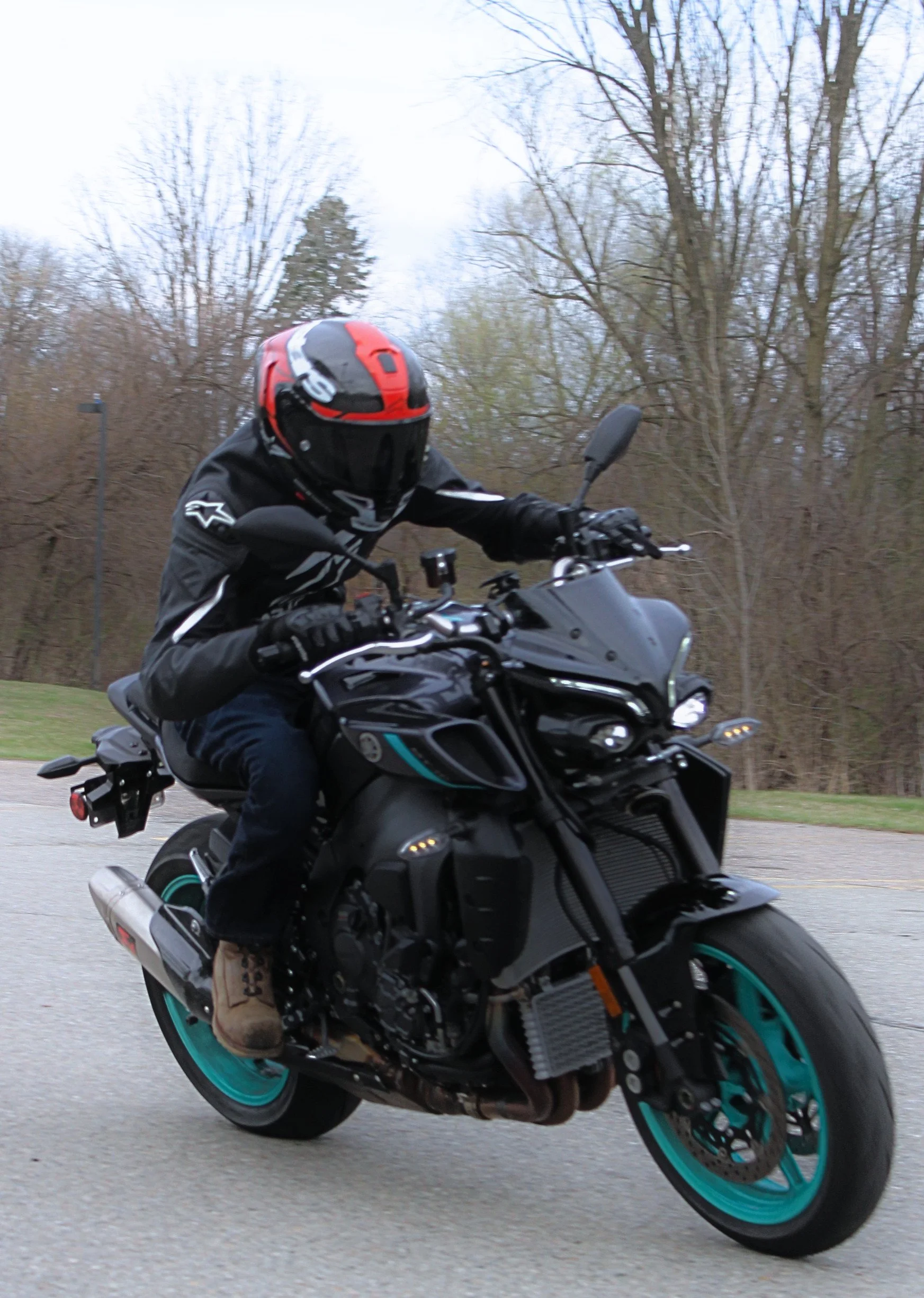 Motorcyclist riding a black motorcycle on a road, wearing a black helmet with red accents, a black jacket, and brown boots, with a leafless tree and cloudy sky in the background.