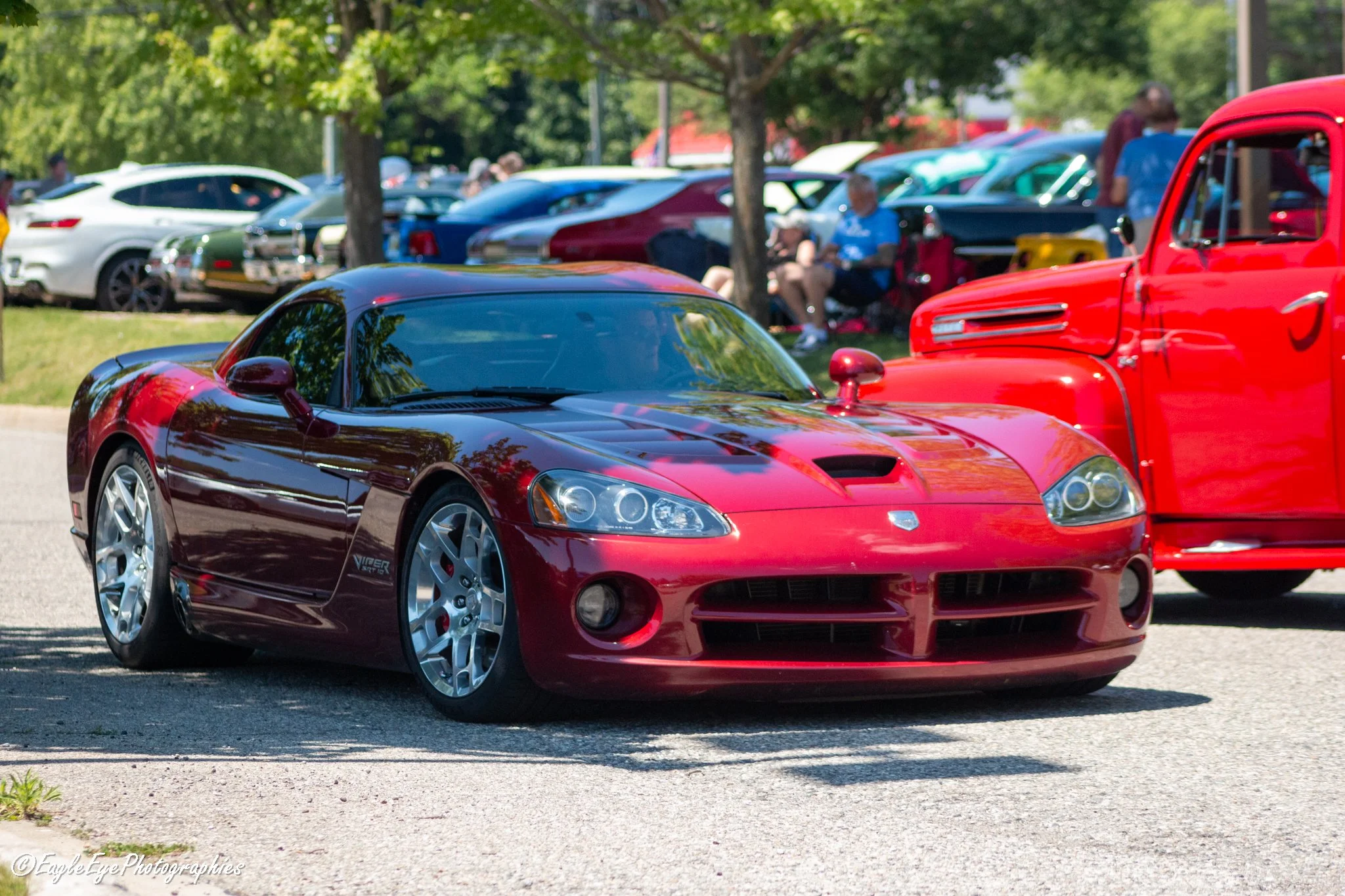 A red sports car parked in a lot during a car show with other vehicles and people in the background, shaded by trees.