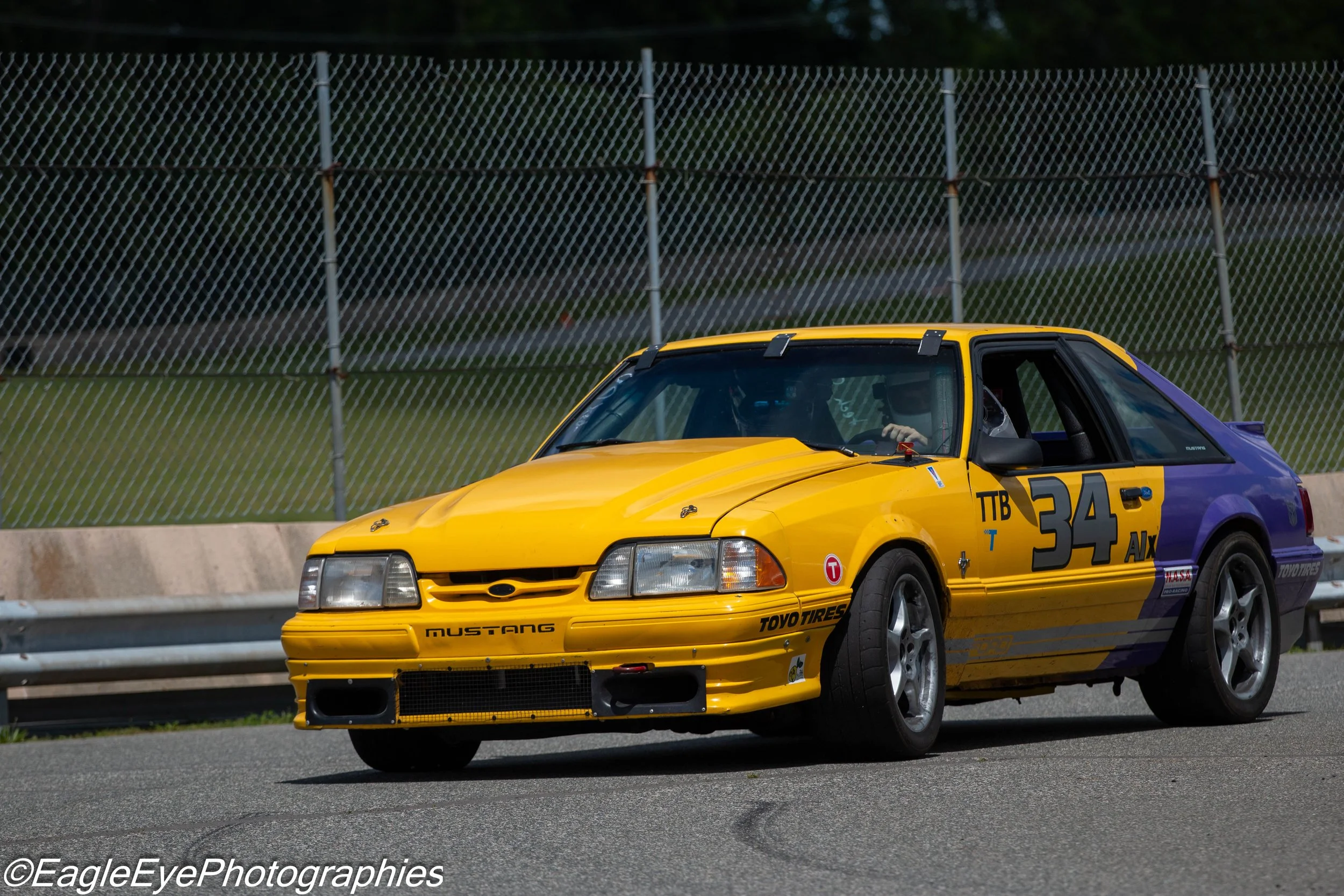 Yellow and purple race car driving on a track with a chain-link fence in the background.