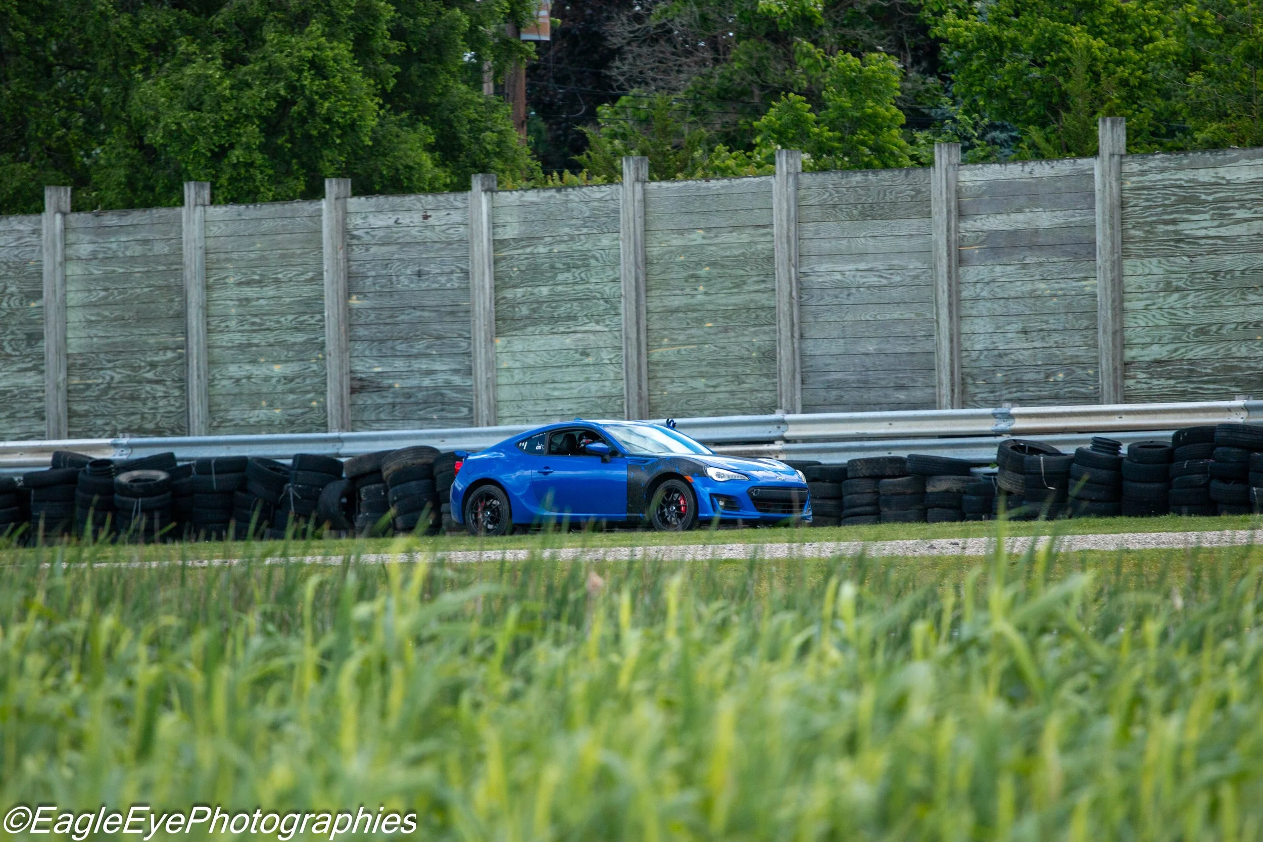 A blue sports car on a race track behind tires and a tall gray wooden fence, with green trees in the background.