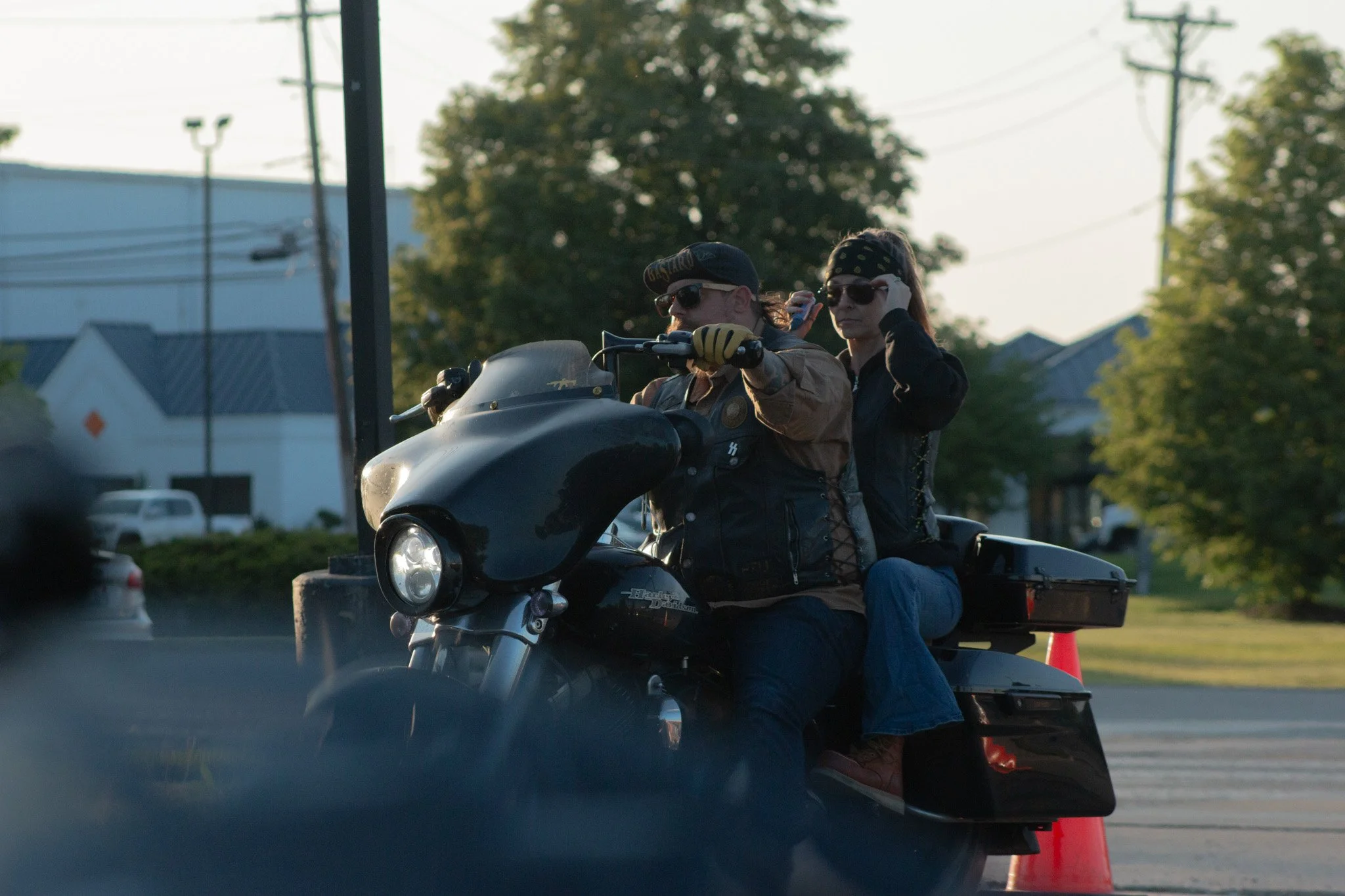 Two motorcyclists riding on a Harley Davidson motorcycle on a road with trees and power lines in the background during daylight.