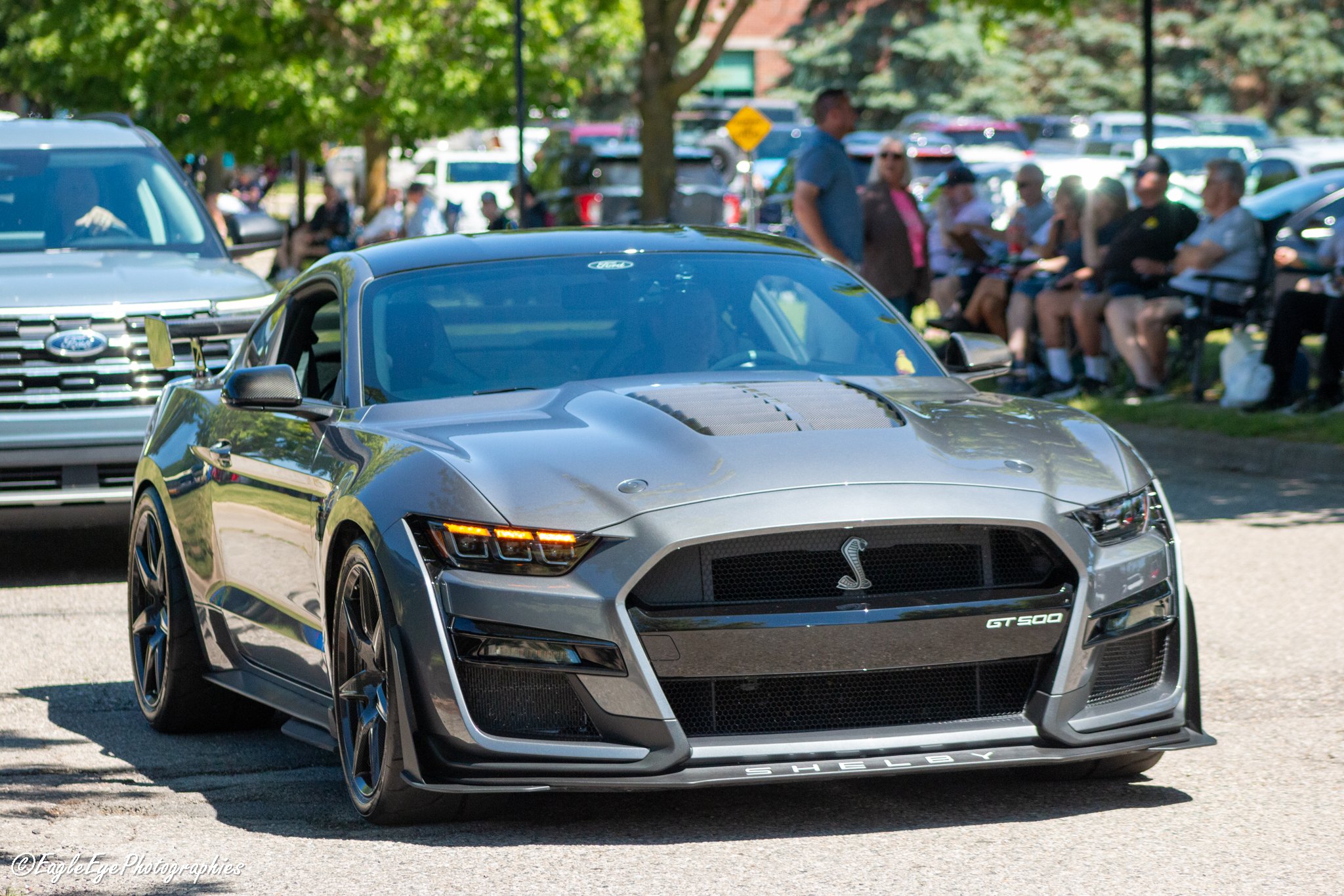 A silver Ford Shelby GT500 sports car parked outdoors with a group of people sitting on benches and standing in the background, surrounded by trees and other parked cars.