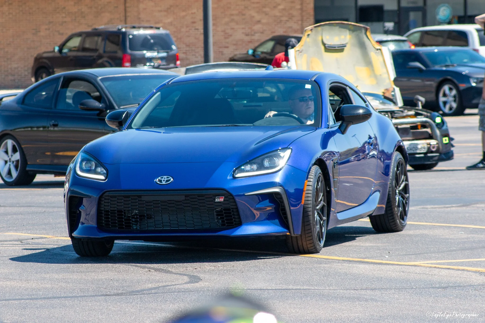 A blue Toyota sports car in a parking lot during daytime, with a person driving it. Several other cars are parked in the background near a brick building.
