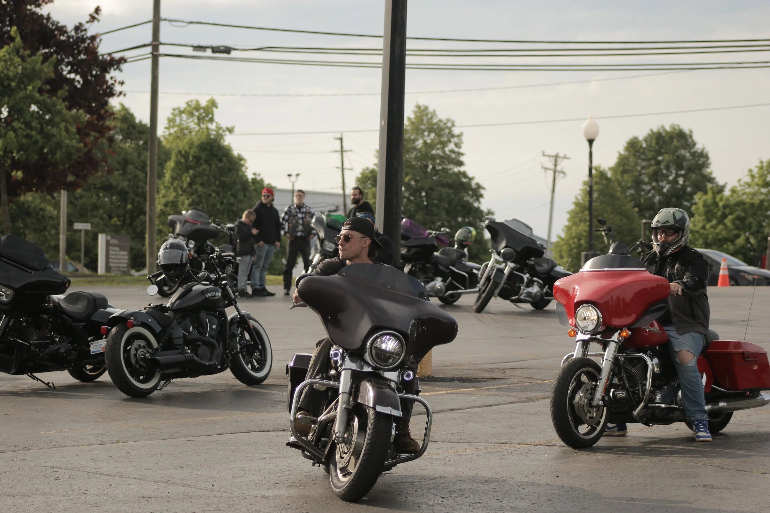 People gathered around motorcycles and a sidecar in a parking lot, with green trees and power lines in the background.