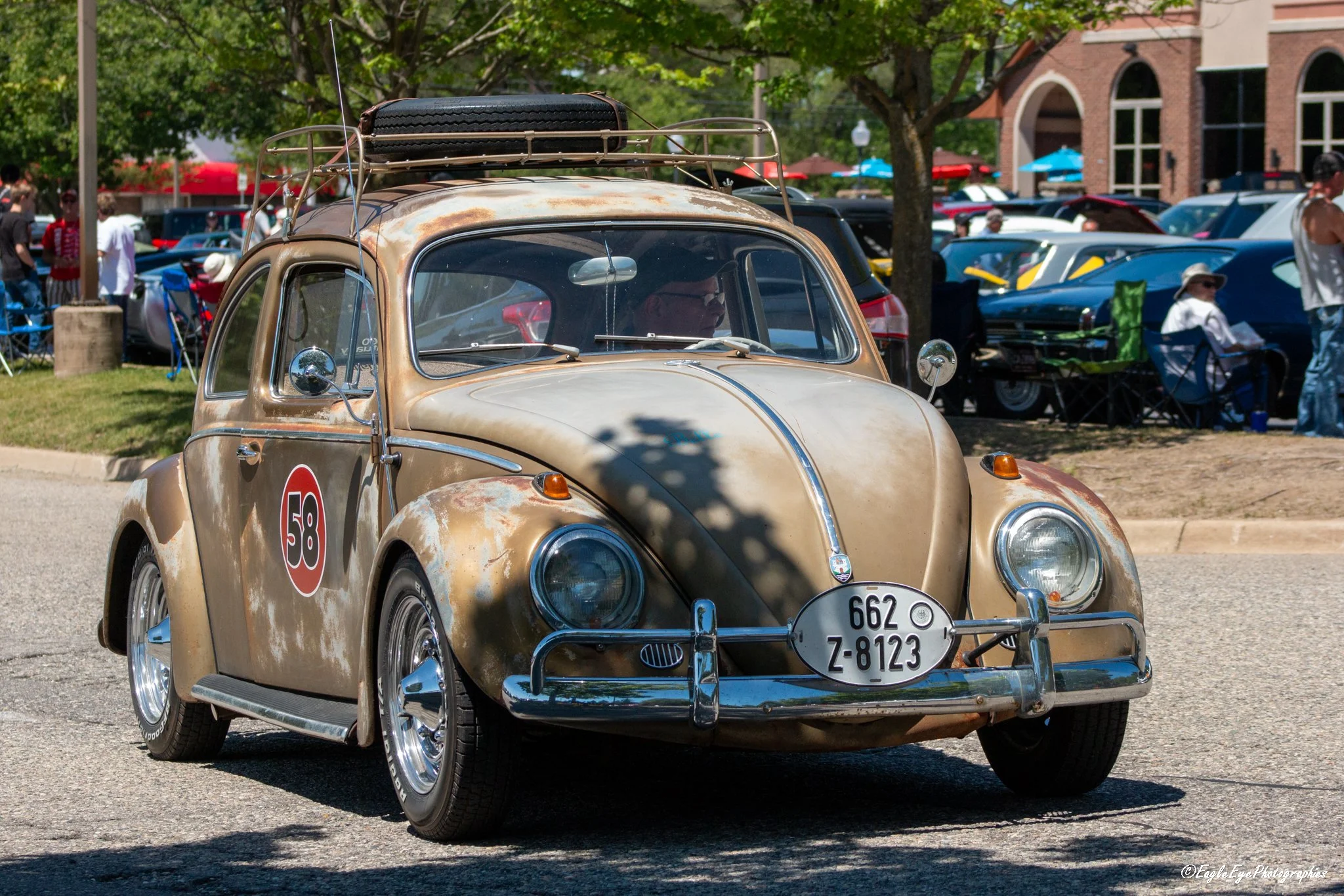 Vintage small beige car with rust spots and racing number 58 on the side parked at an outdoor car show with people and trees in the background.