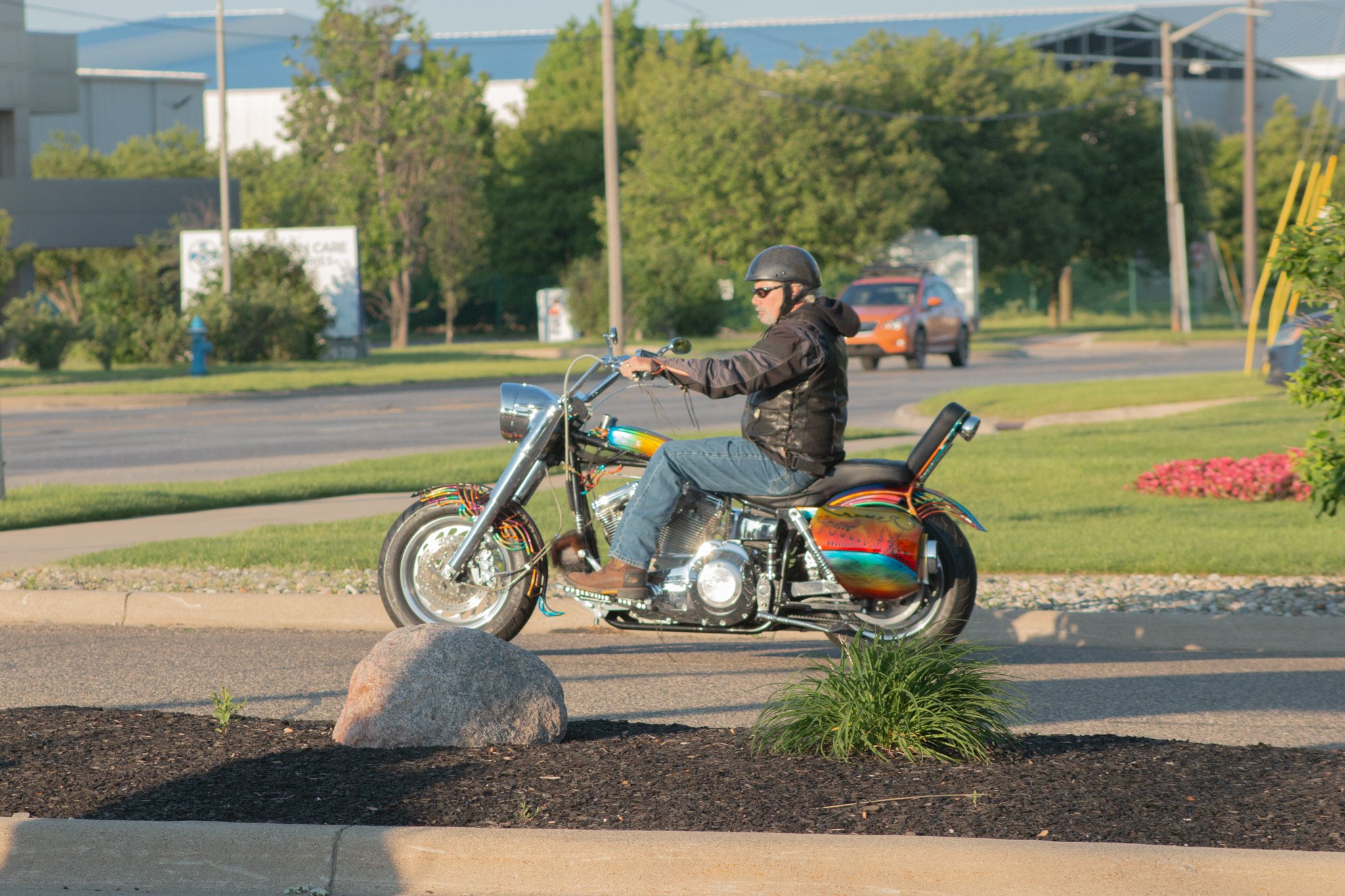 A man riding a custom motorcycle on a street, wearing a black helmet, black jacket, sunglasses, and jeans; the motorcycle has rainbow-colored flames and chrome details, with greenery and buildings in the background.