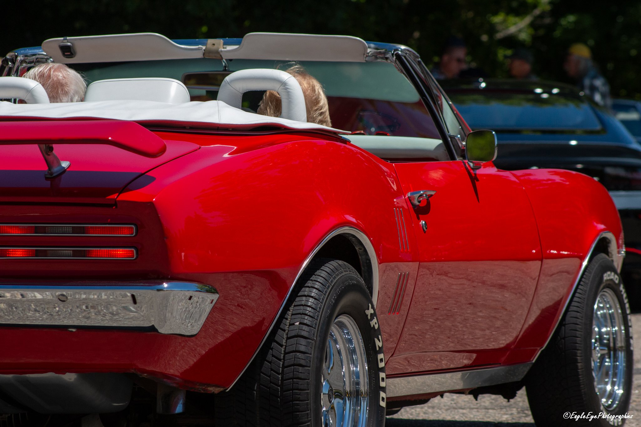A red vintage convertible car with white interior and a white soft top, parked outdoors, with people and trees in the background.