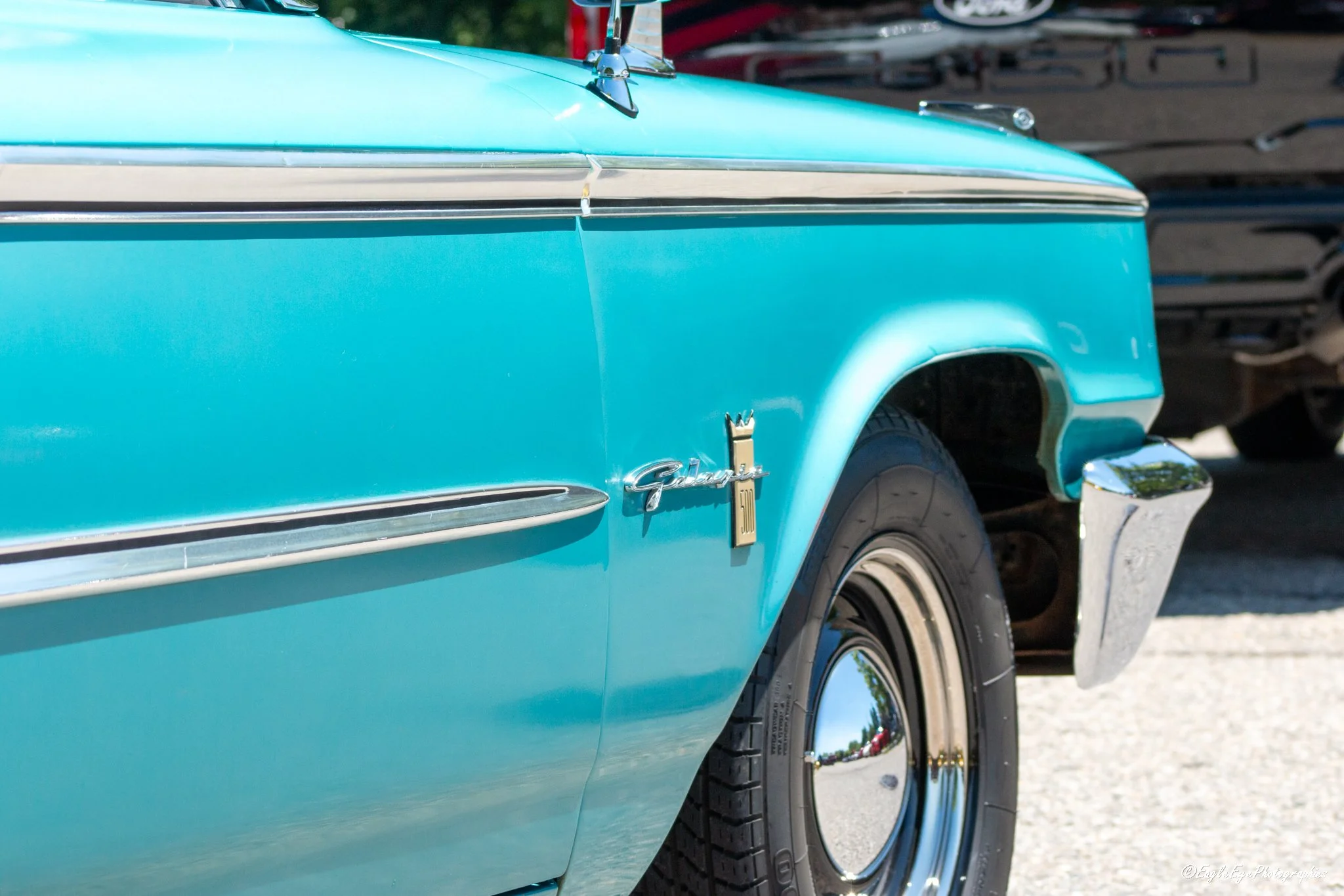 Close-up of the front side of a vintage blue Chevrolet car showing the wheel, part of the body, and chrome details with a blurred background of what appears to be another vehicle and outdoor setting.