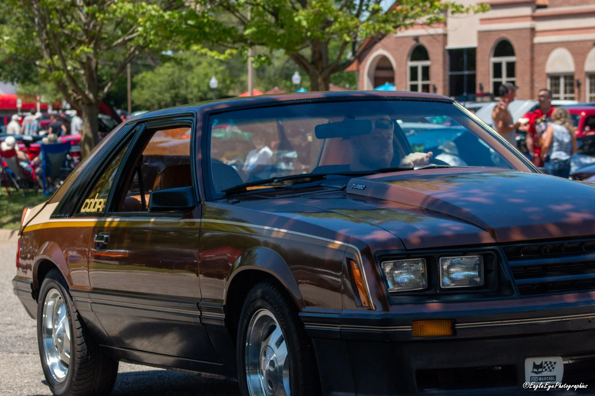 A vintage brown and black car with a Turbo badge on the front, parked outdoors during a car show event with people and trees in the background.