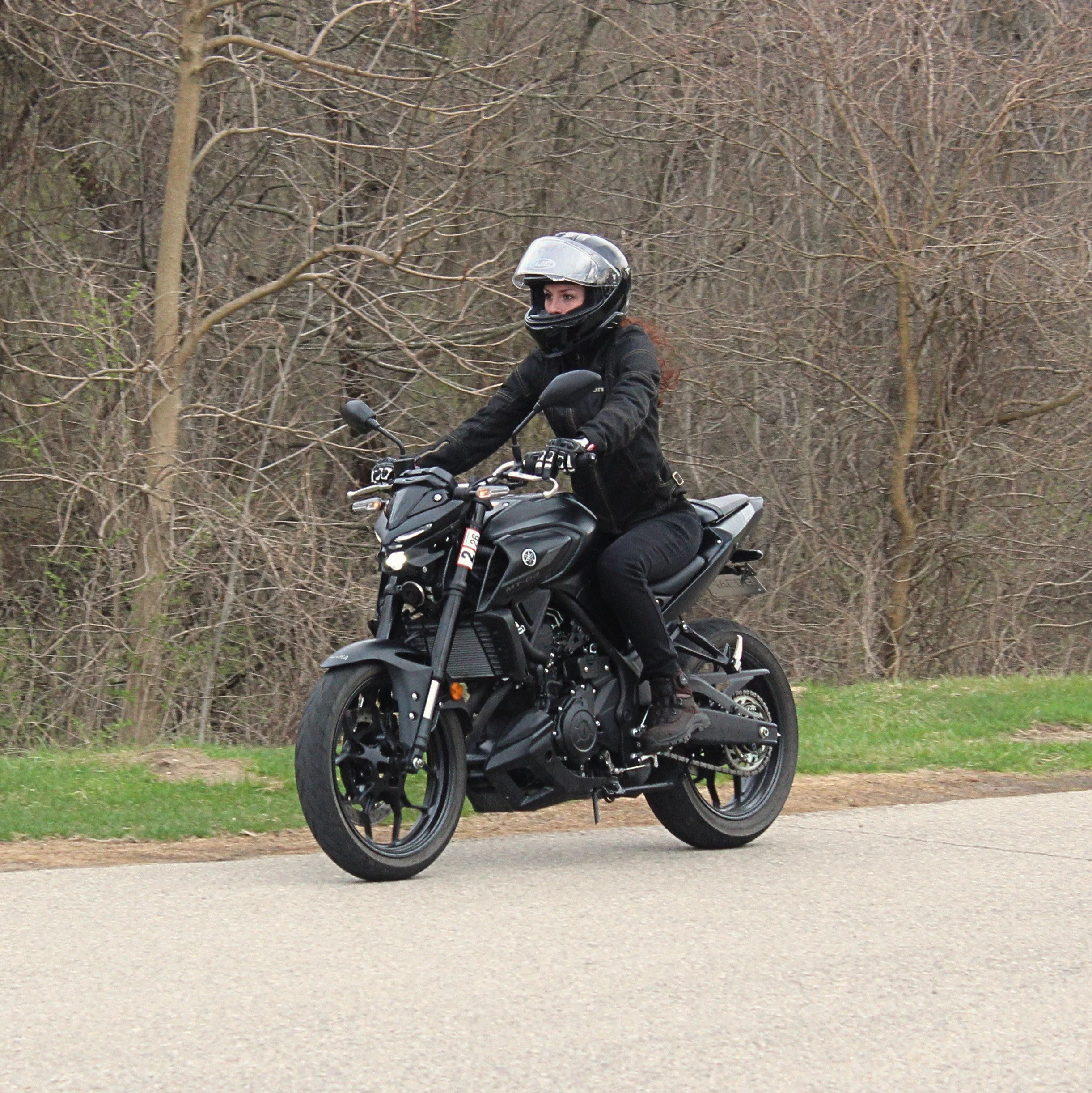 A person wearing a black motorcycle helmet and black riding gear is riding a black motorcycle on a paved road with grass on the side. The background consists of leafless trees and bushes.