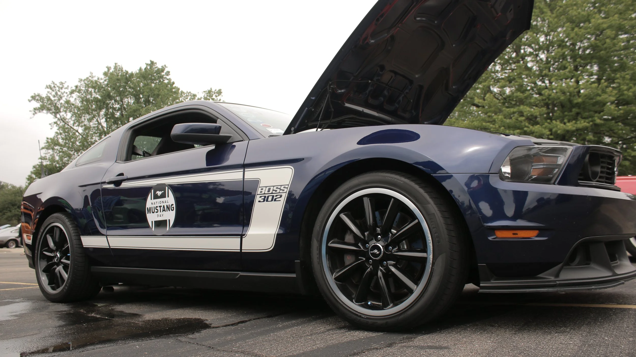 A blue Ford Mustang with its hood open, displaying the engine. The car has a National Mustang Day sticker on the door and the model name Boss 302. It is parked outdoors on a cloudy day with trees in the background.