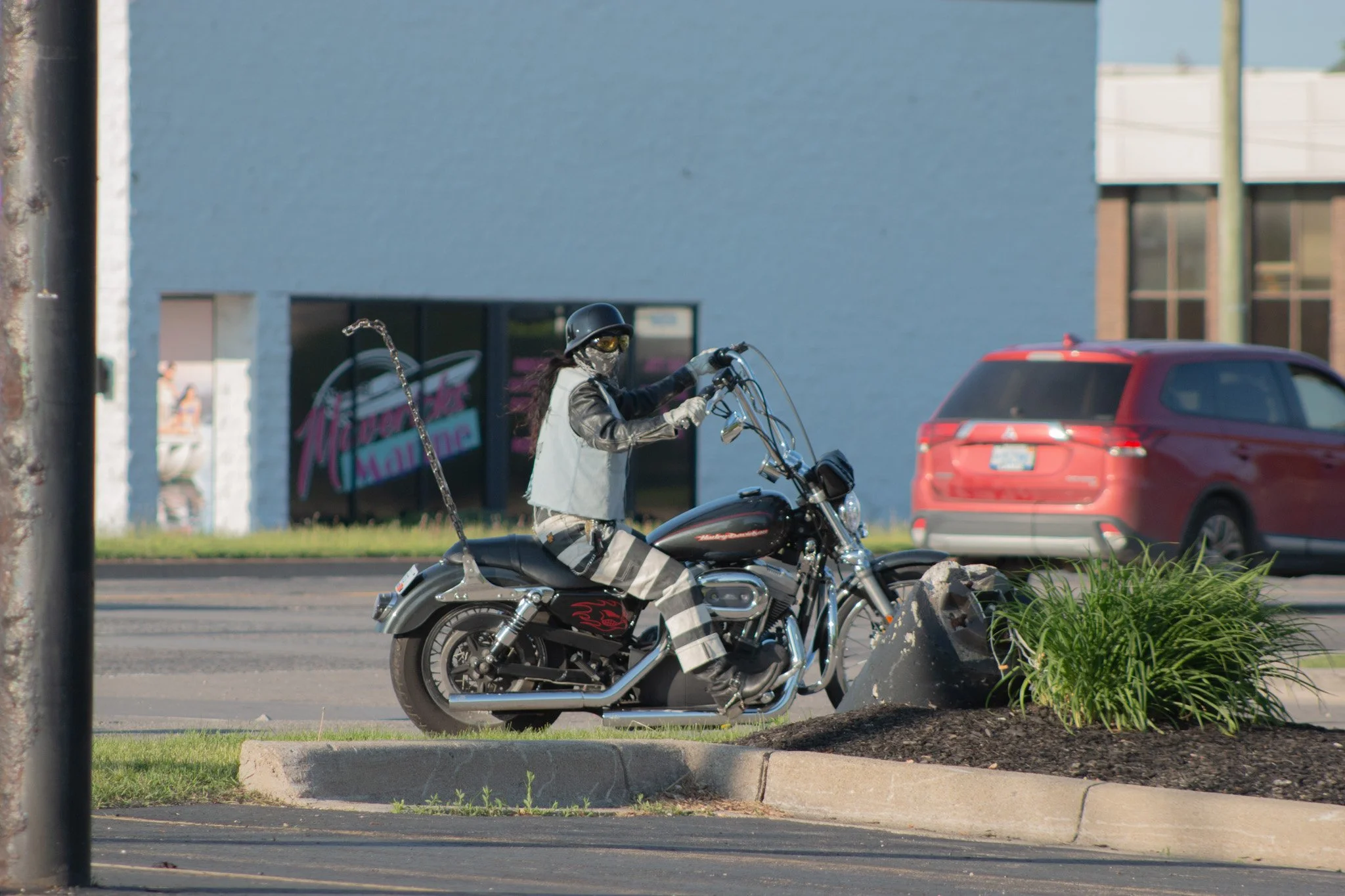 A woman wearing a motorcycle helmet, face mask, and protective clothing riding a black Harley-Davidson motorcycle on the street, with a red car and a commercial building in the background.
